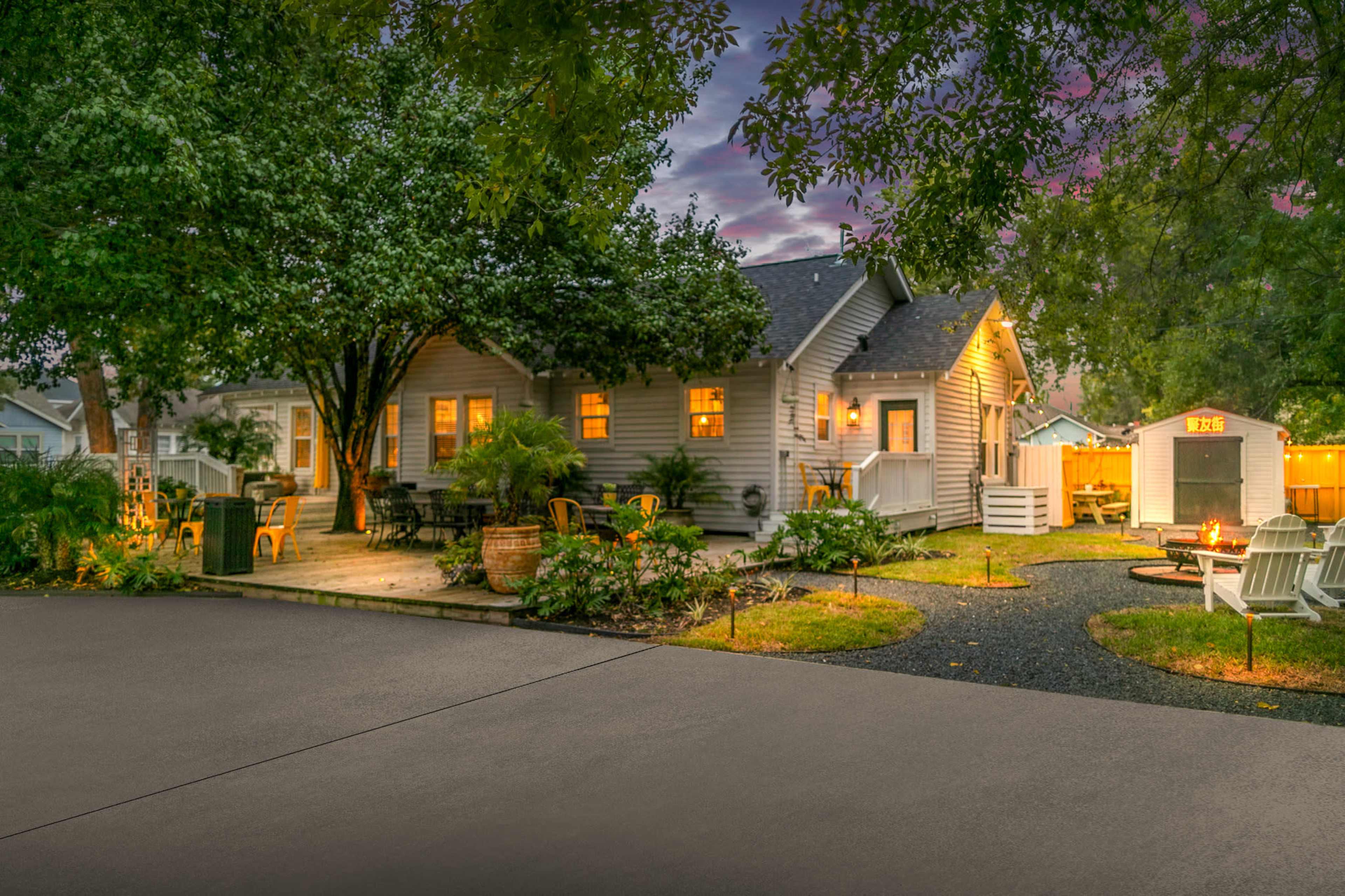 A well-lit backyard with a wooden house, outdoor seating, and a fire pit surrounded by lush greenery and garden lights at dusk.