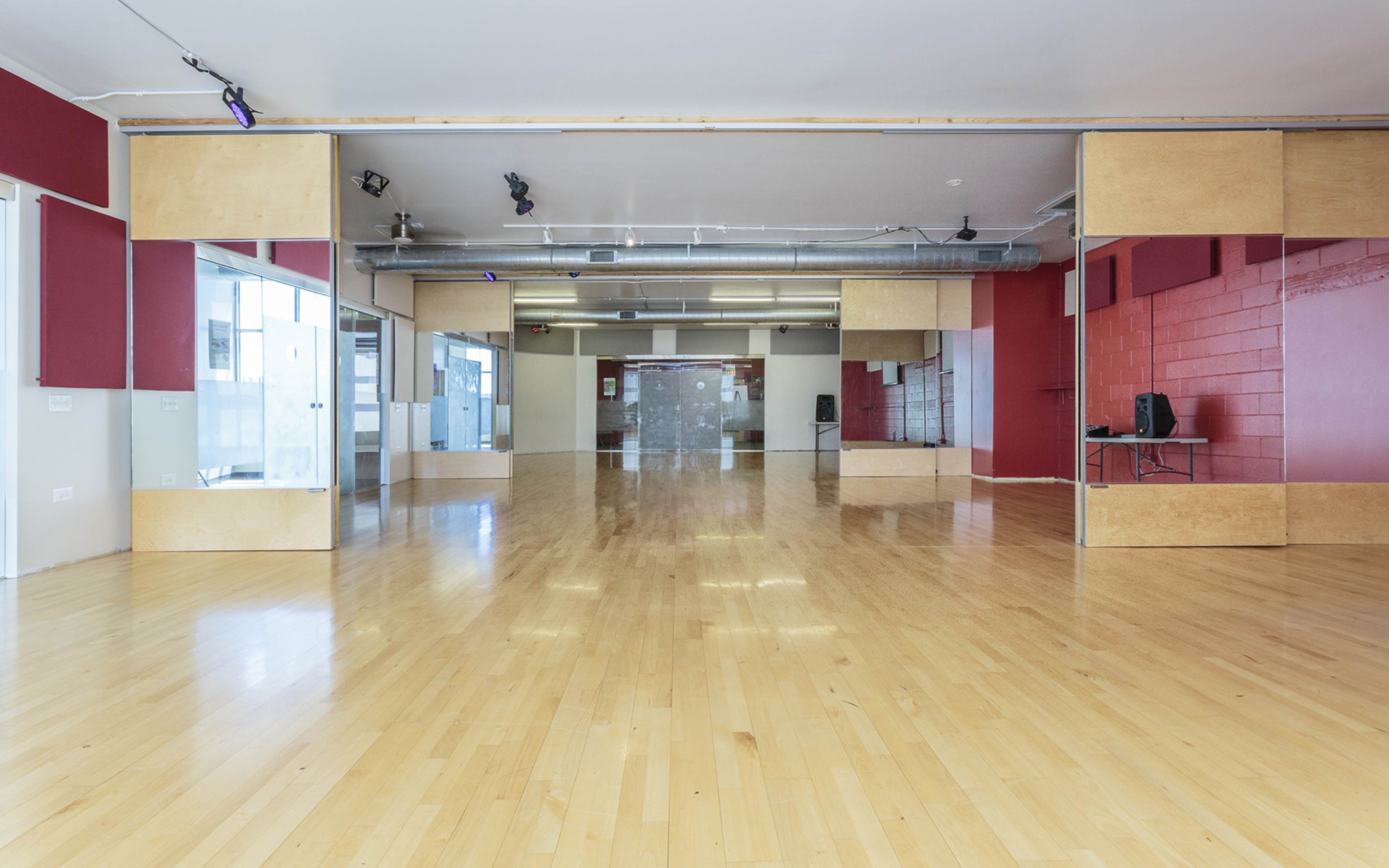 The image shows a spacious, empty studio with a wooden floor, large mirrors on one wall, and red accents on the walls.