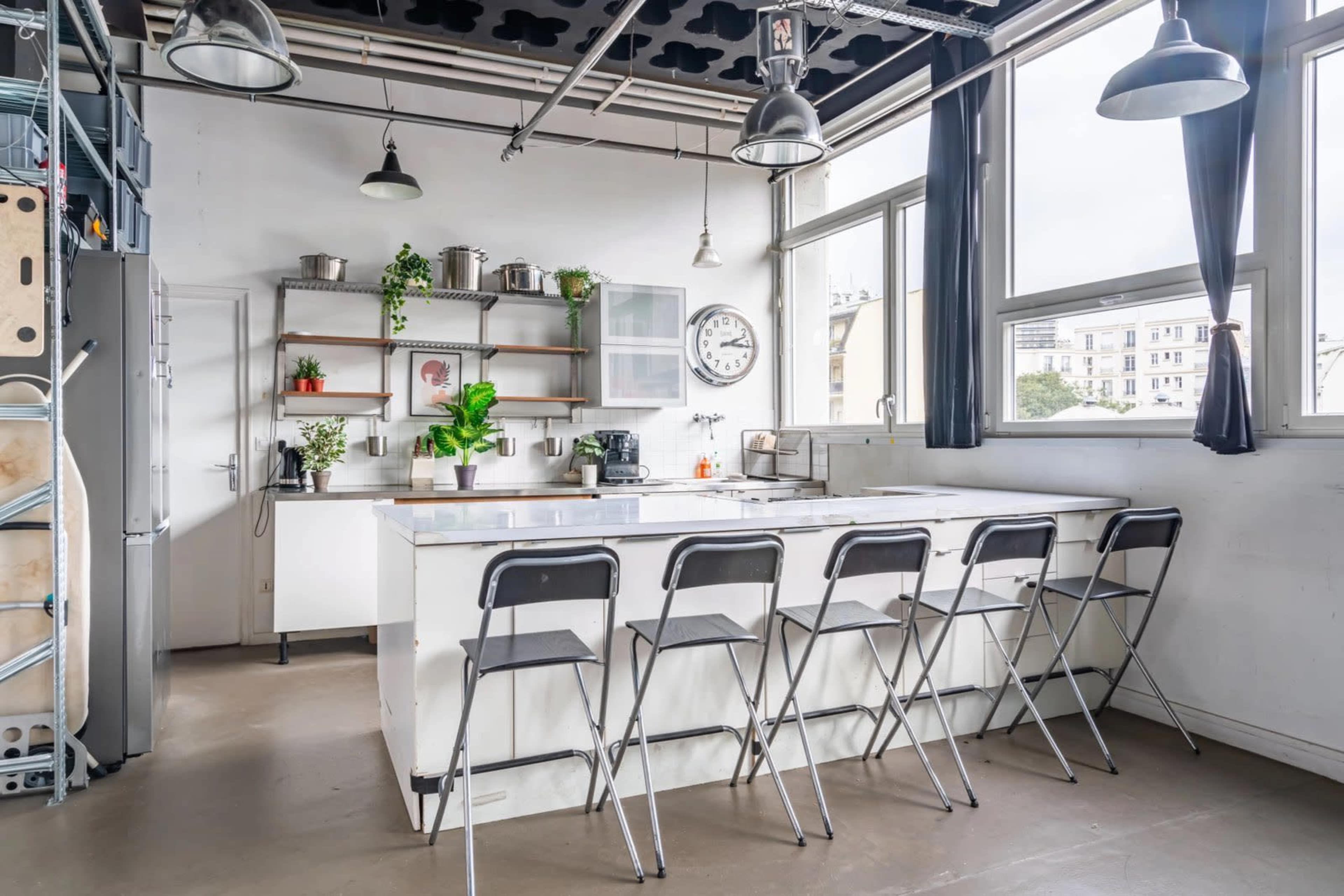 The image shows a modern kitchen with a white island, metal stools, open shelves, and large windows allowing natural light.