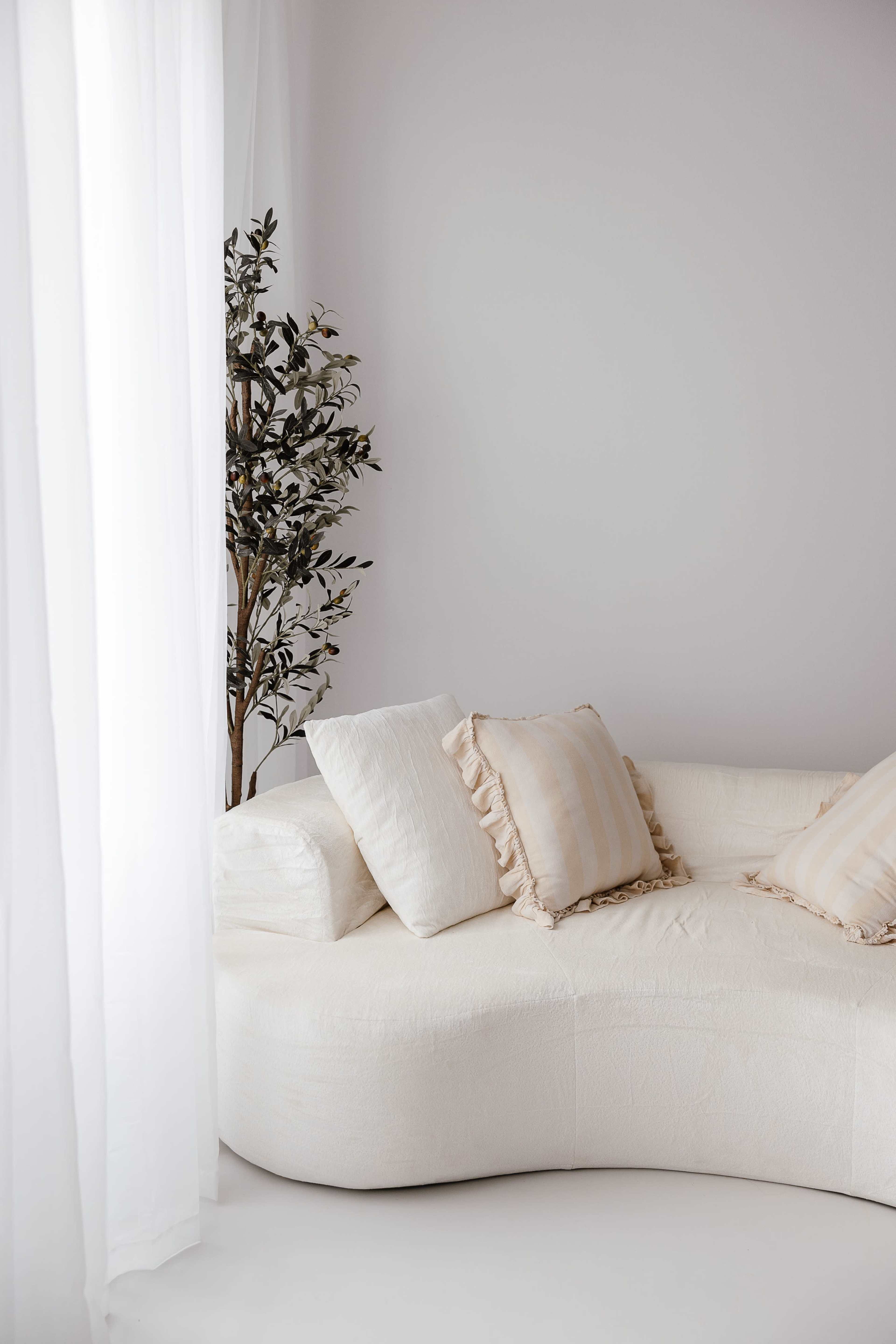 A curved, white sofa with two decorative pillows is positioned next to a potted plant, near sheer white curtains.