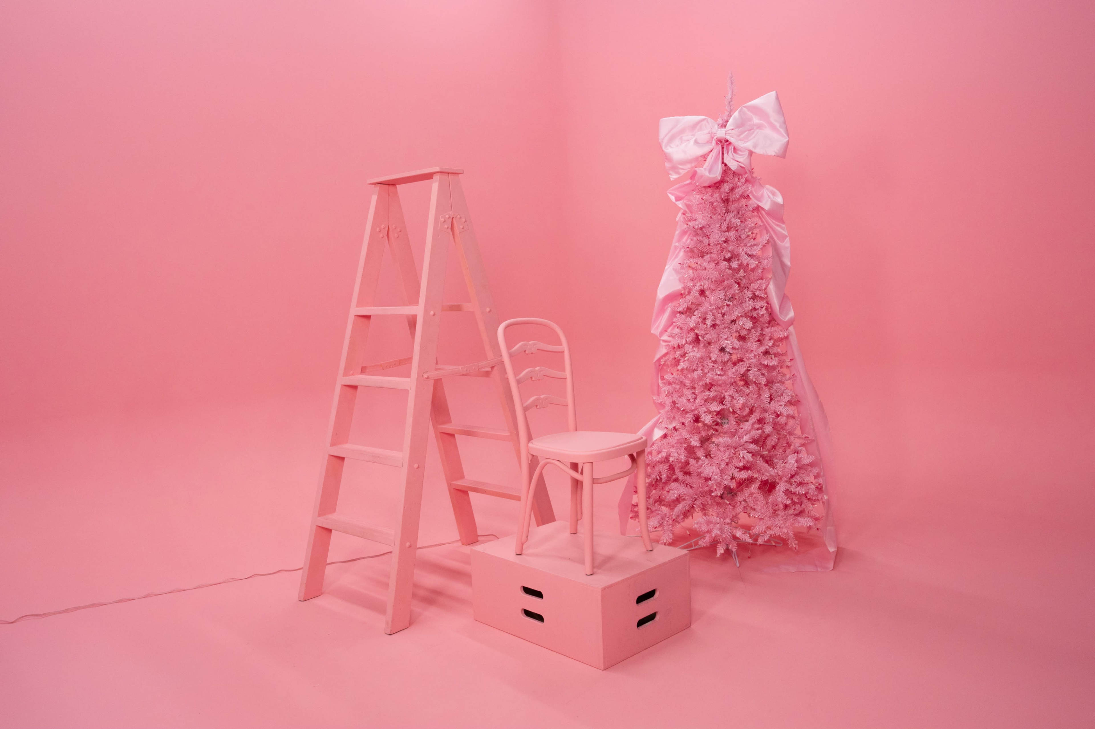 The scene features a pink-themed setup with a pink ladder, a pink chair, and a pink Christmas tree adorned with ribbons, all against a matching pink background.