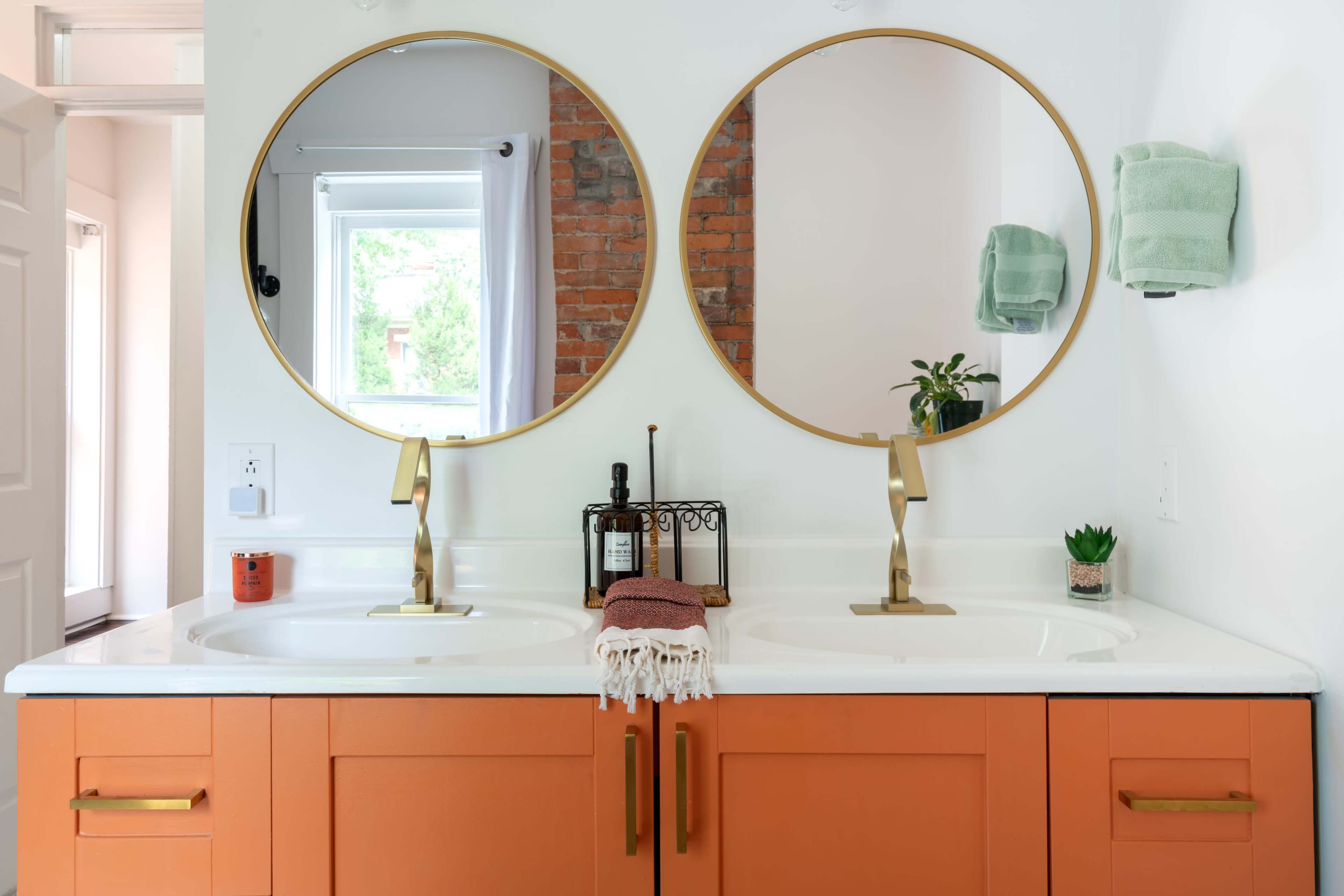 A bright bathroom features an orange double vanity with two round mirrors and a window, complemented by green towels and decorative plants.