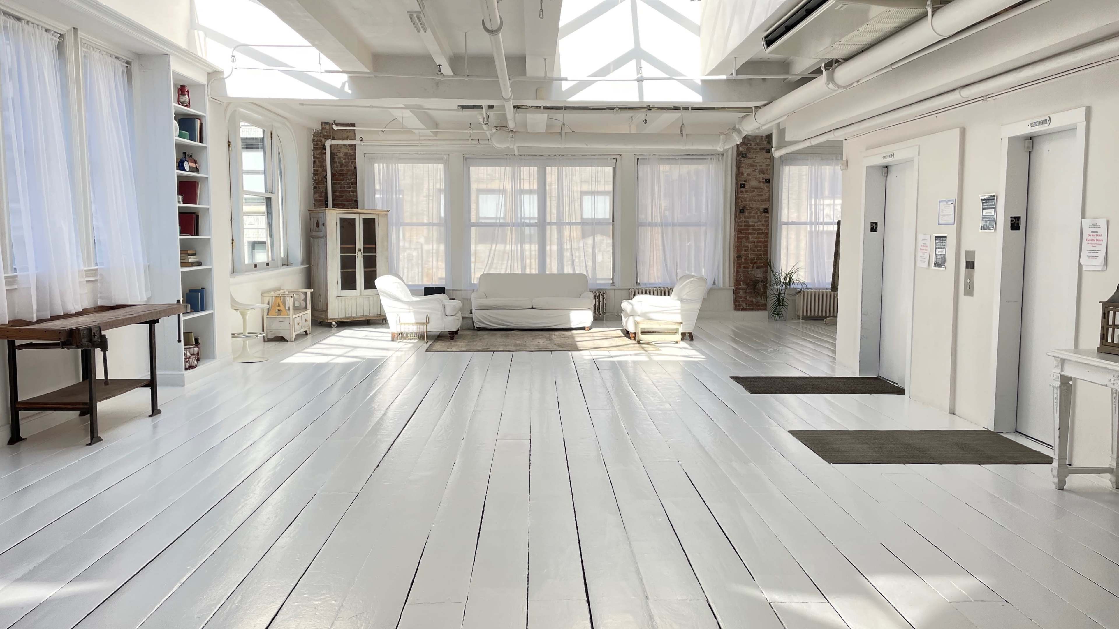 A bright, open space with white wooden floors, large windows, and minimalist furniture including a sofa and chairs.