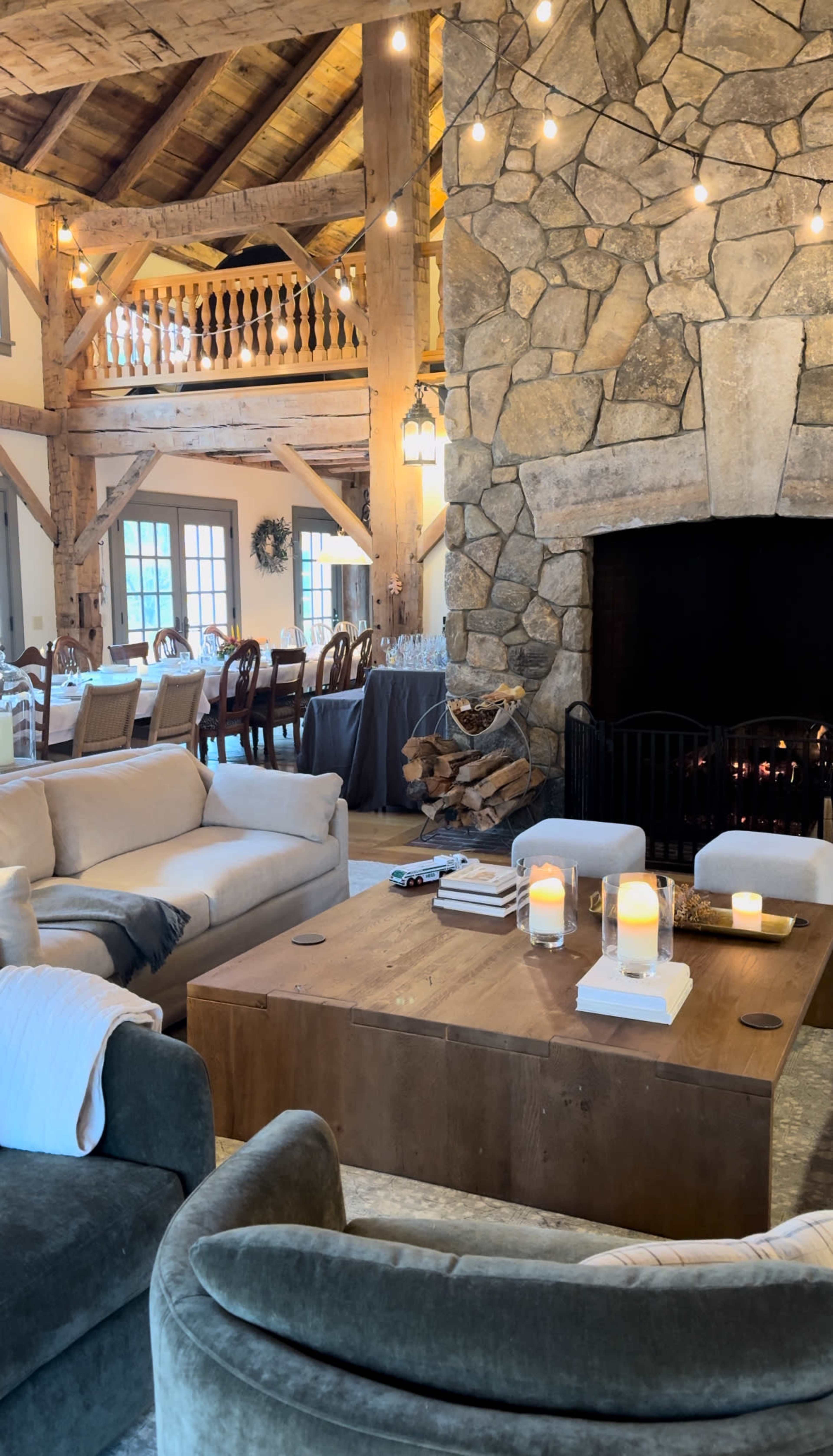 The image shows a cozy interior with a stone fireplace, wooden beams, a large coffee table, and comfortable seating arranged in a spacious room with dining tables in the background.