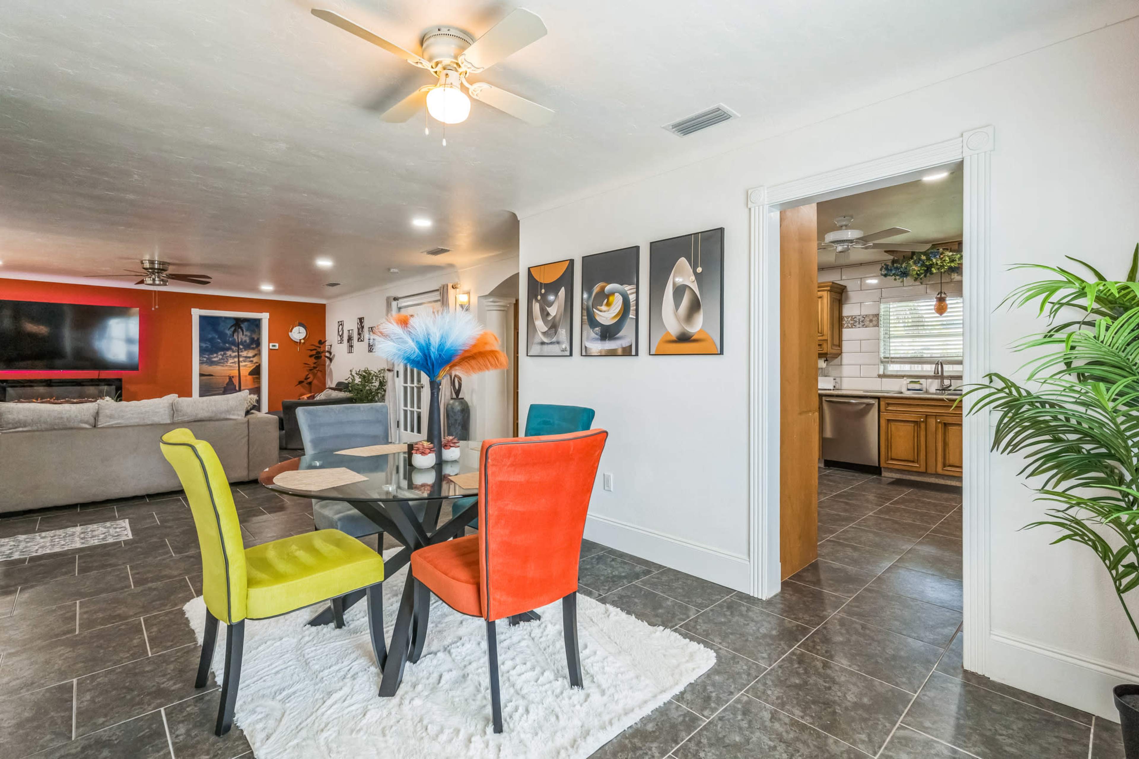 The image shows a modern living area with a glass dining table surrounded by colorful chairs, a plush rug, and a view into a kitchen area with wooden cabinetry.
