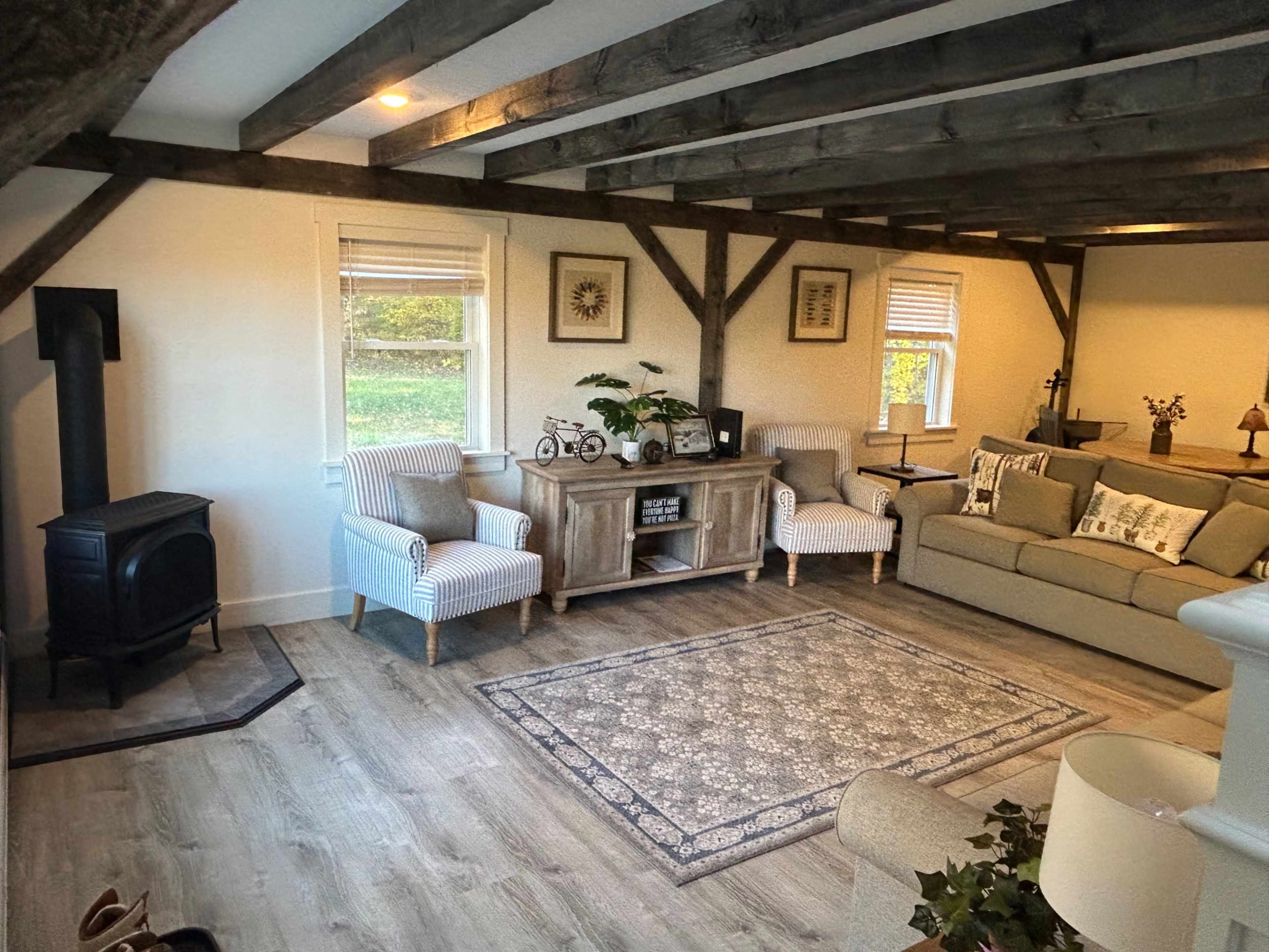 The living room features a beige sectional sofa, two striped chairs, a wooden coffee table, and a small black wood stove beneath exposed wooden beams.