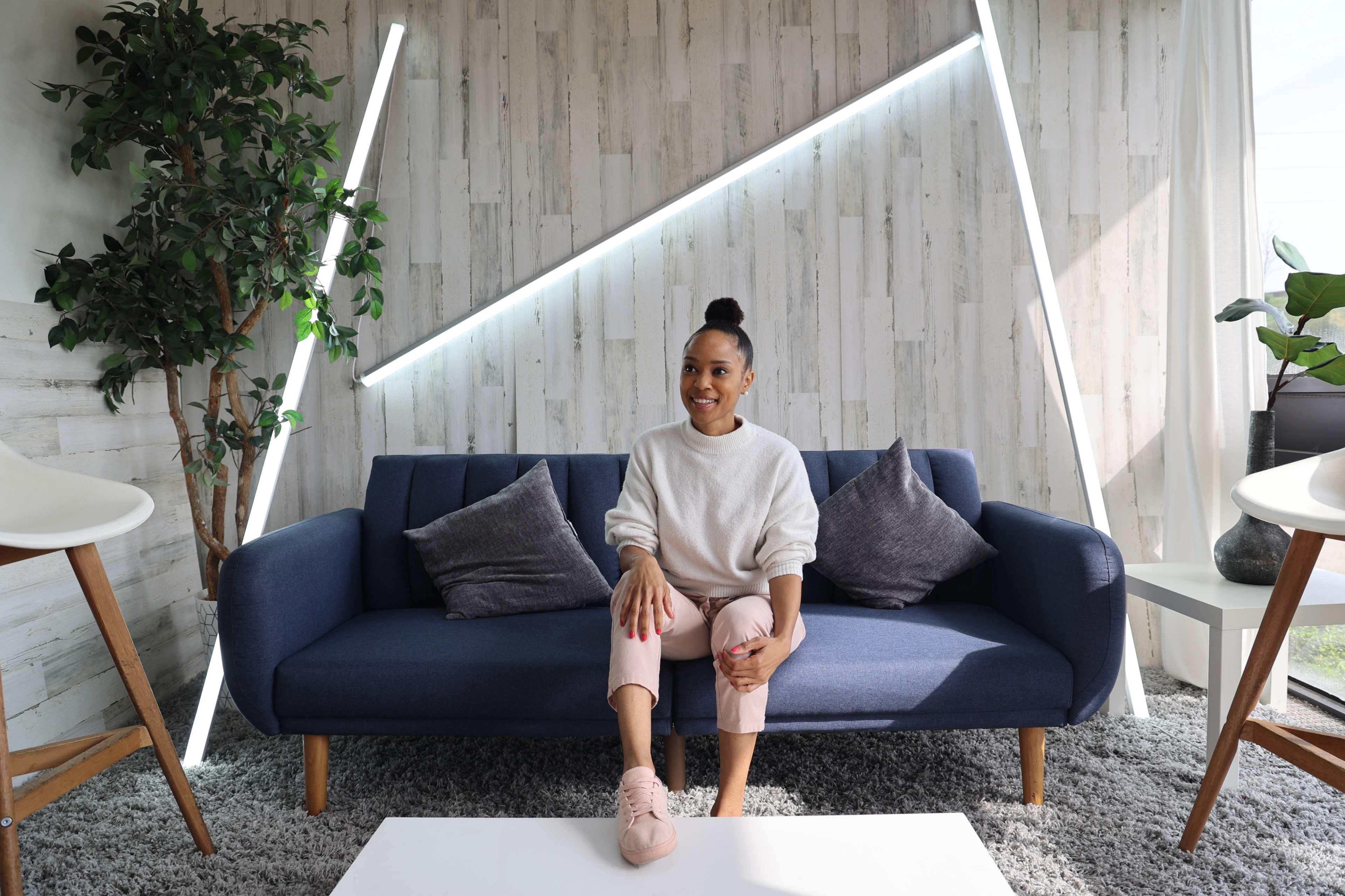 A woman sits on a blue sofa in a stylishly decorated room with plants and modern lighting.