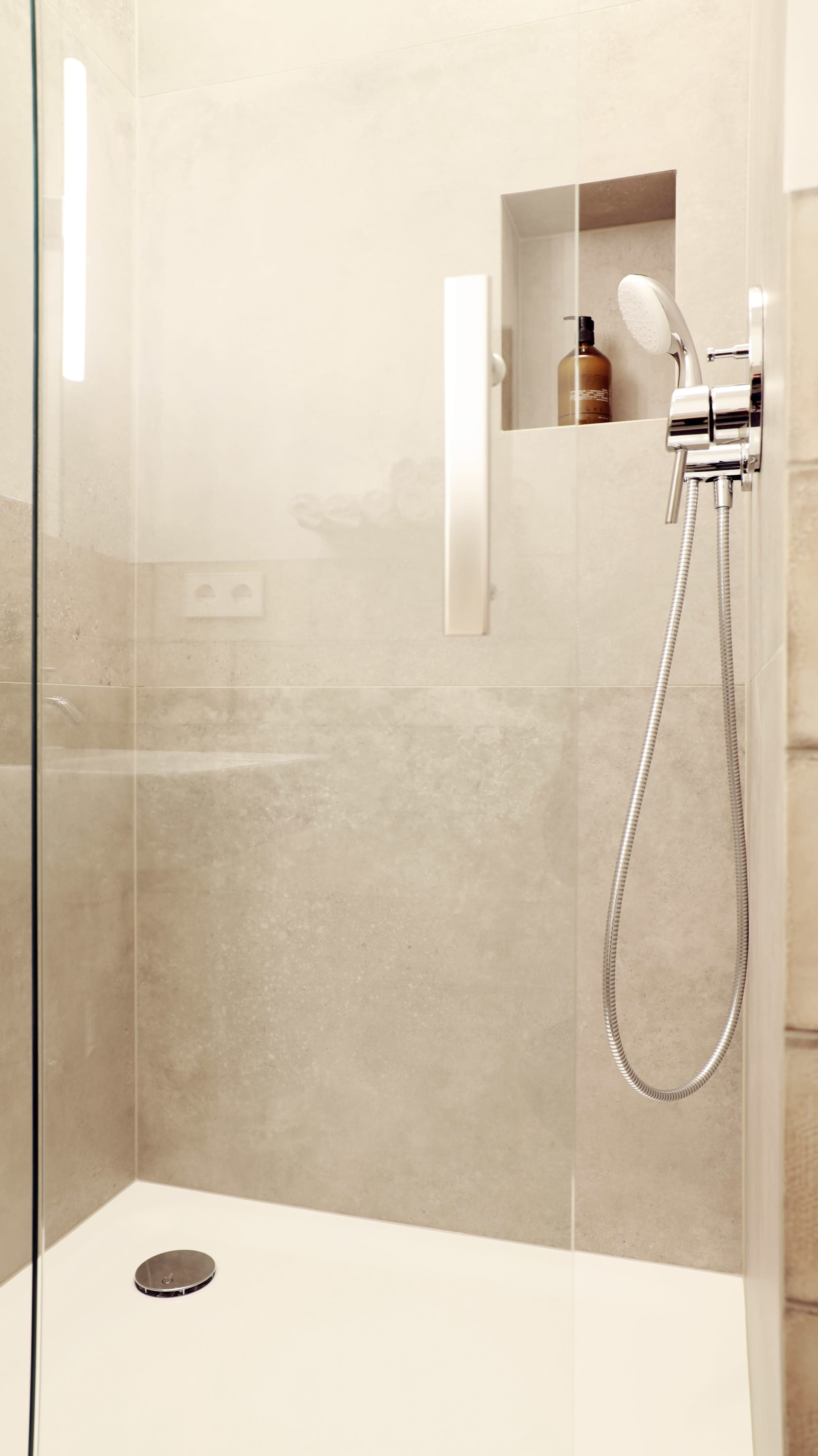 The image shows a modern shower with a glass door, stainless steel showerhead, and light-colored tiled walls.