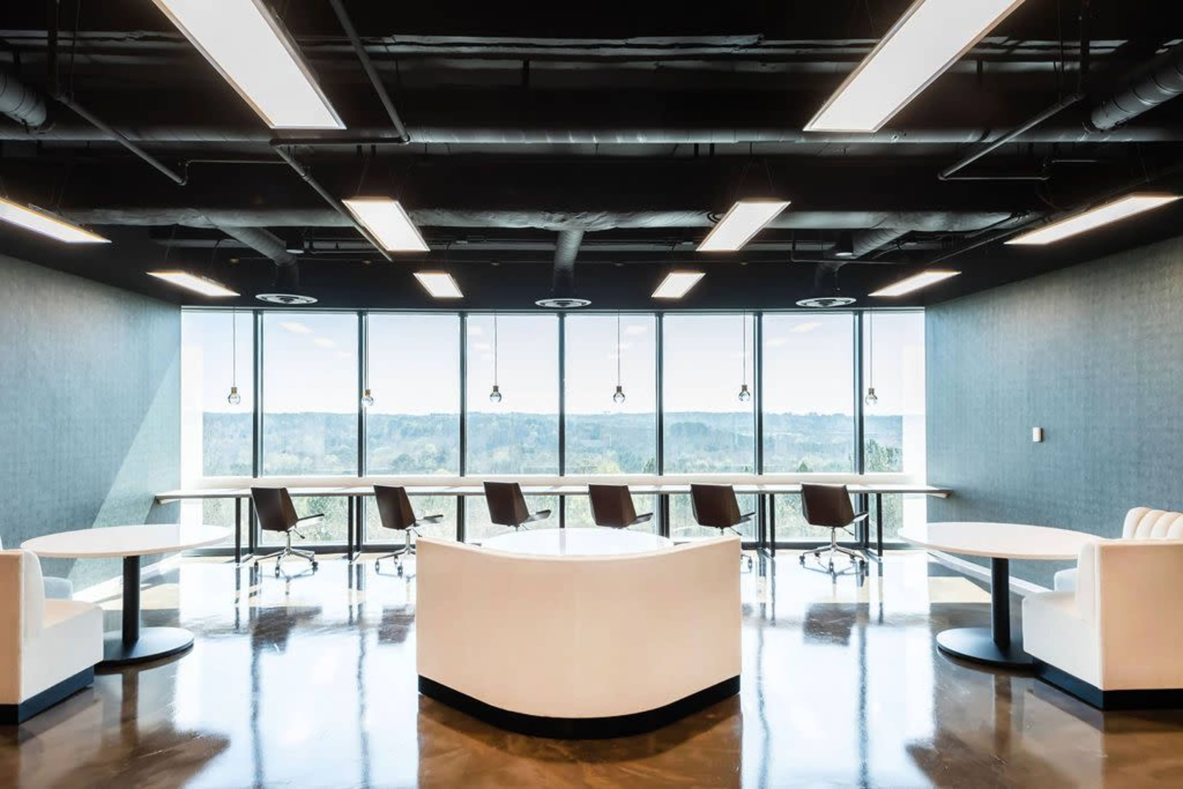 The image shows a modern office space with large windows, featuring several round tables and high-backed chairs arranged in a well-lit setting.