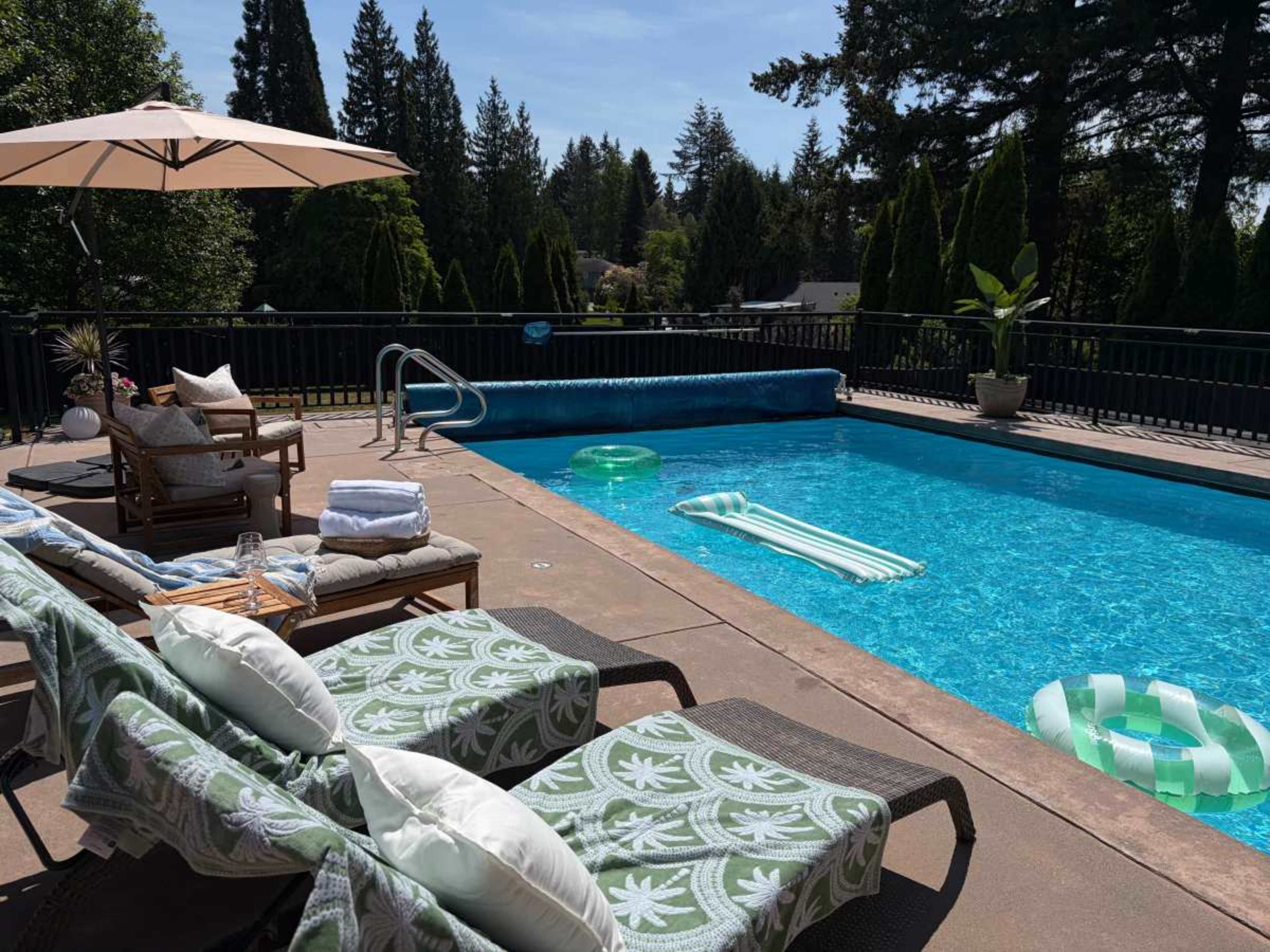 A clear blue swimming pool surrounded by lounge chairs, towels, and inflatable floats, with trees and a patio umbrella in the background.