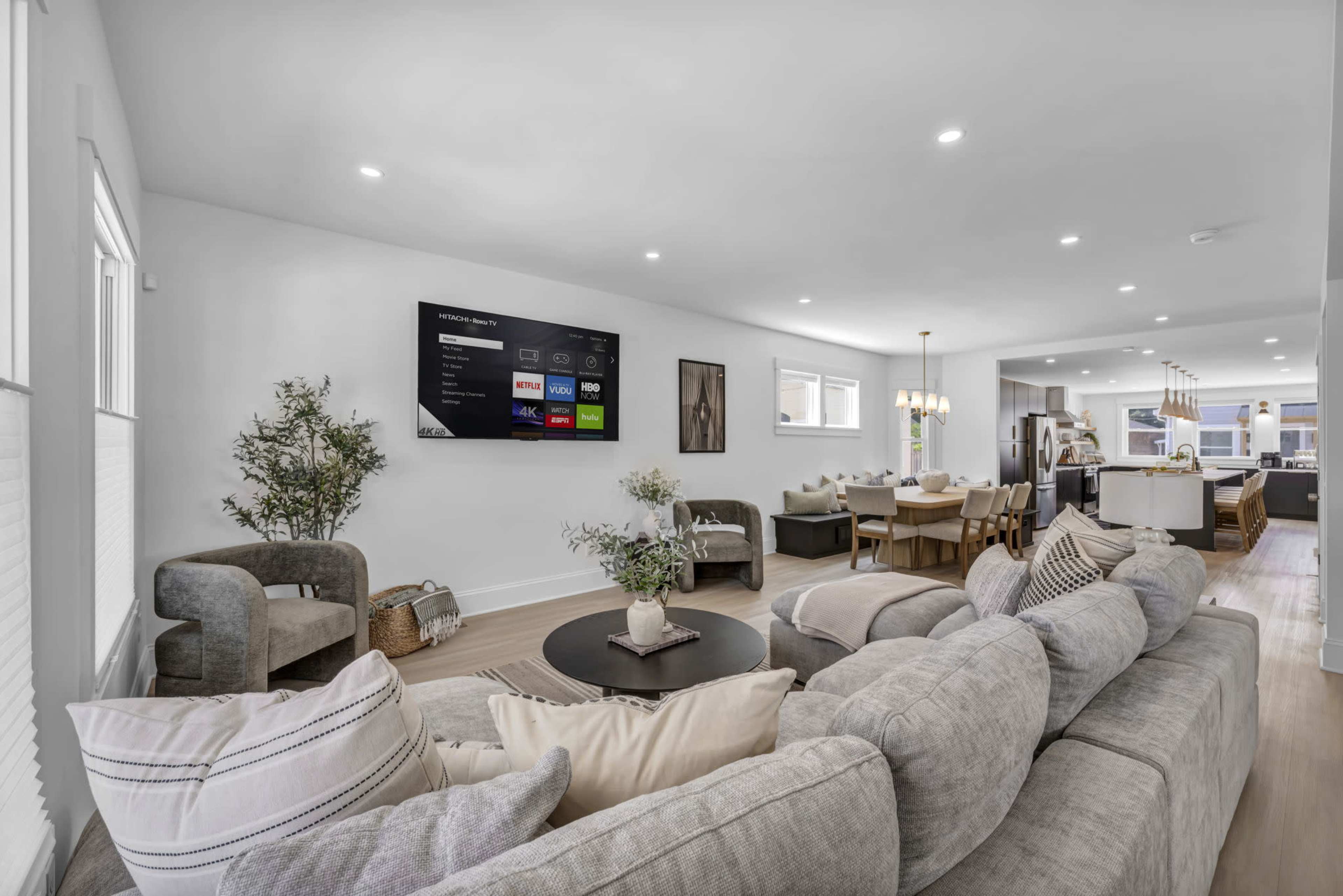 A modern living room features a large sectional sofa, a round coffee table, and a wall-mounted television with a dining area visible in the background.