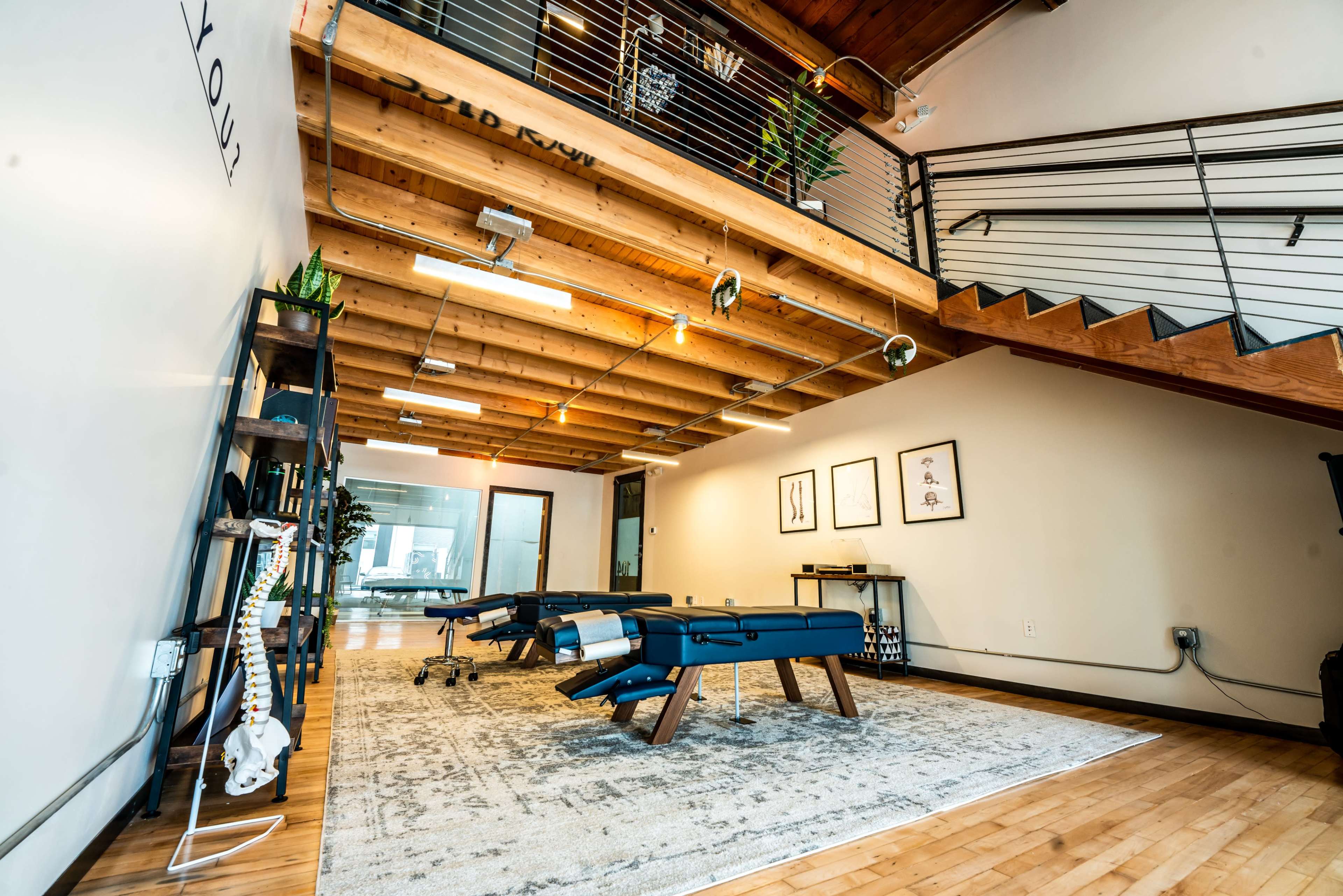 A spacious interior with wooden beams and a carpeted area containing treatment tables and decorative plants, leading to a staircase.