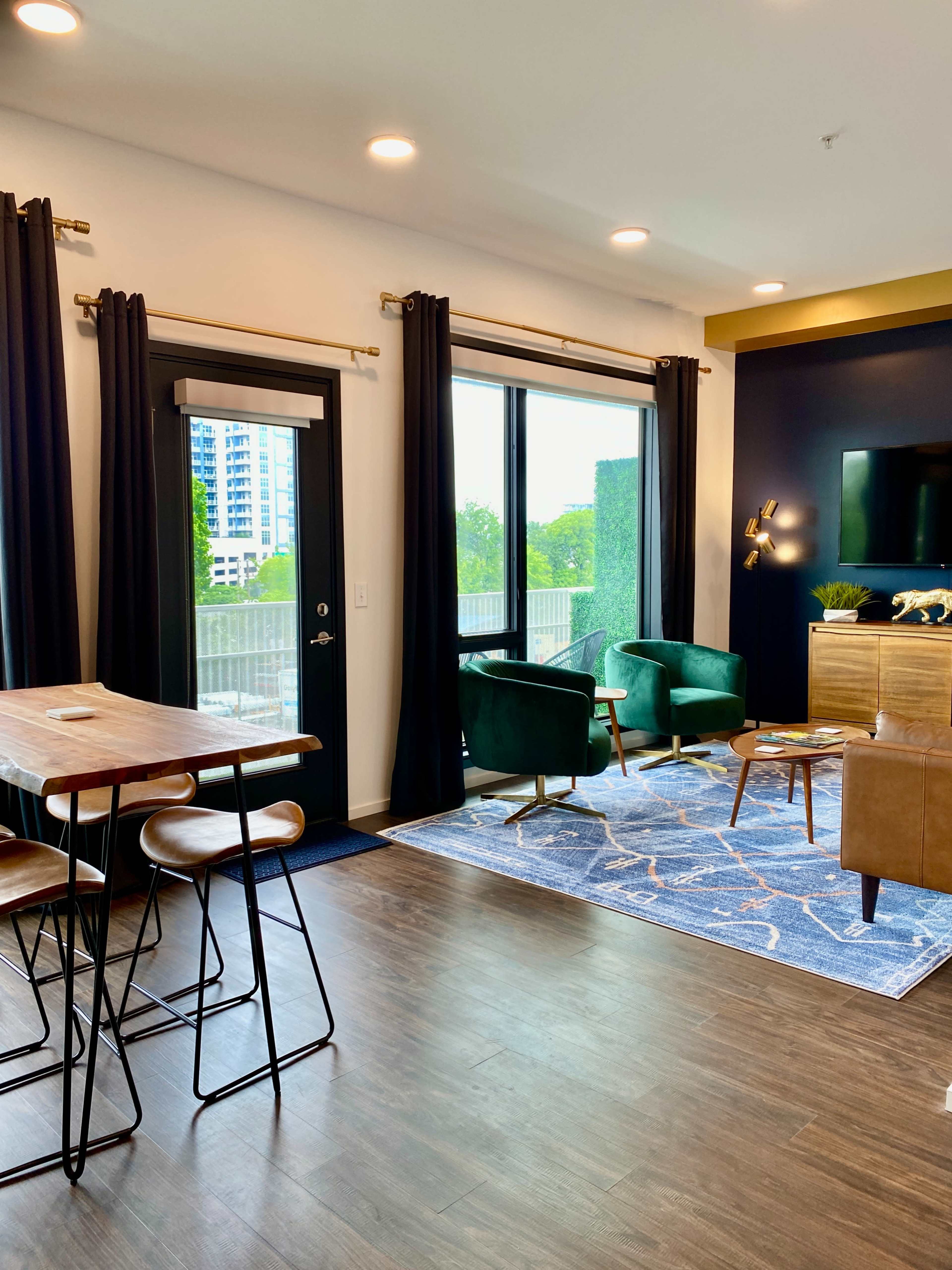 A modern living space featuring a dining table with bar stools, two green armchairs, a brown wooden cabinet, a television, and large windows with dark curtains.