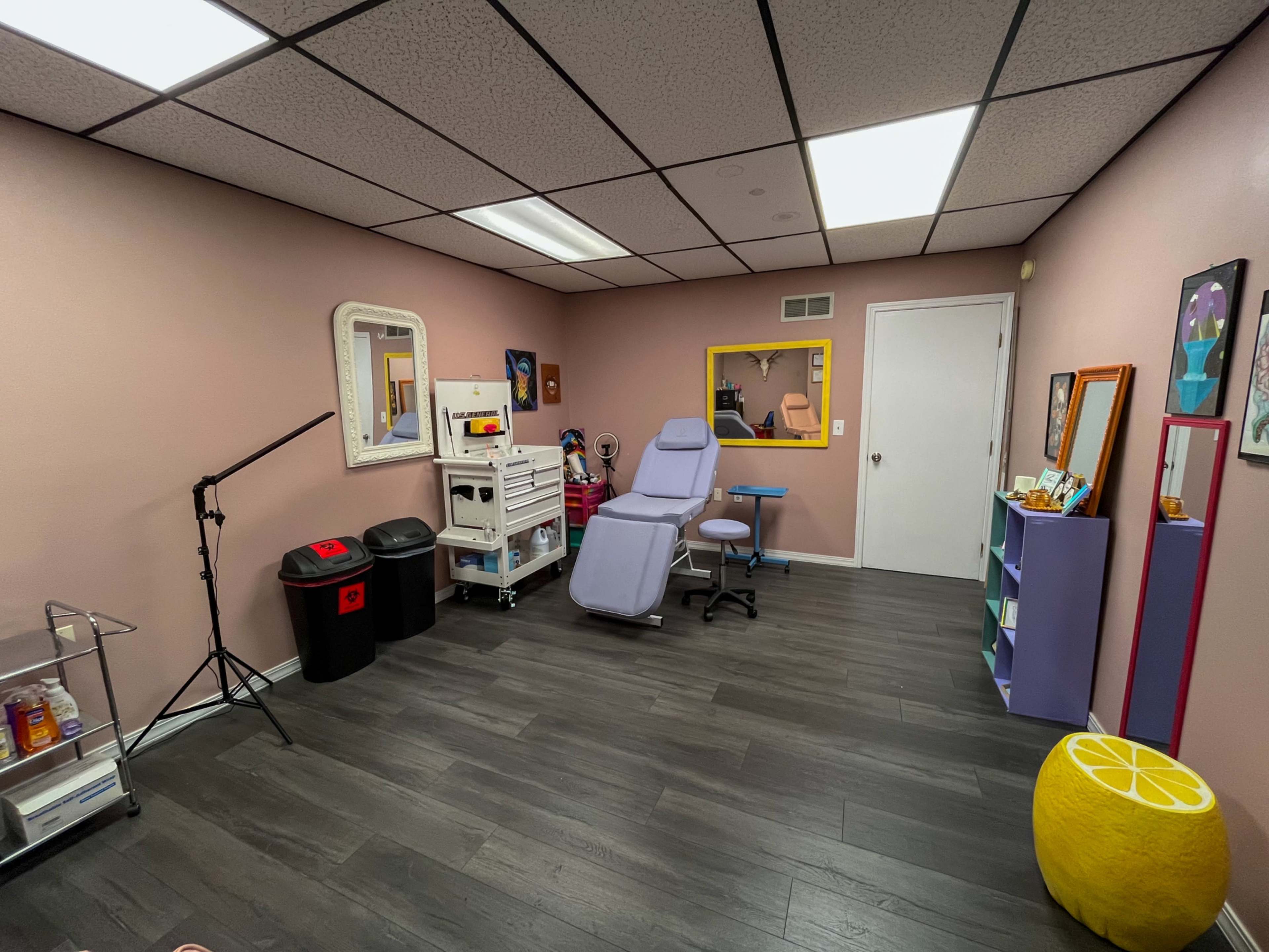 The image shows a small, clinical room containing a treatment chair, medical equipment, and storage units, with pink walls and a gray floor.