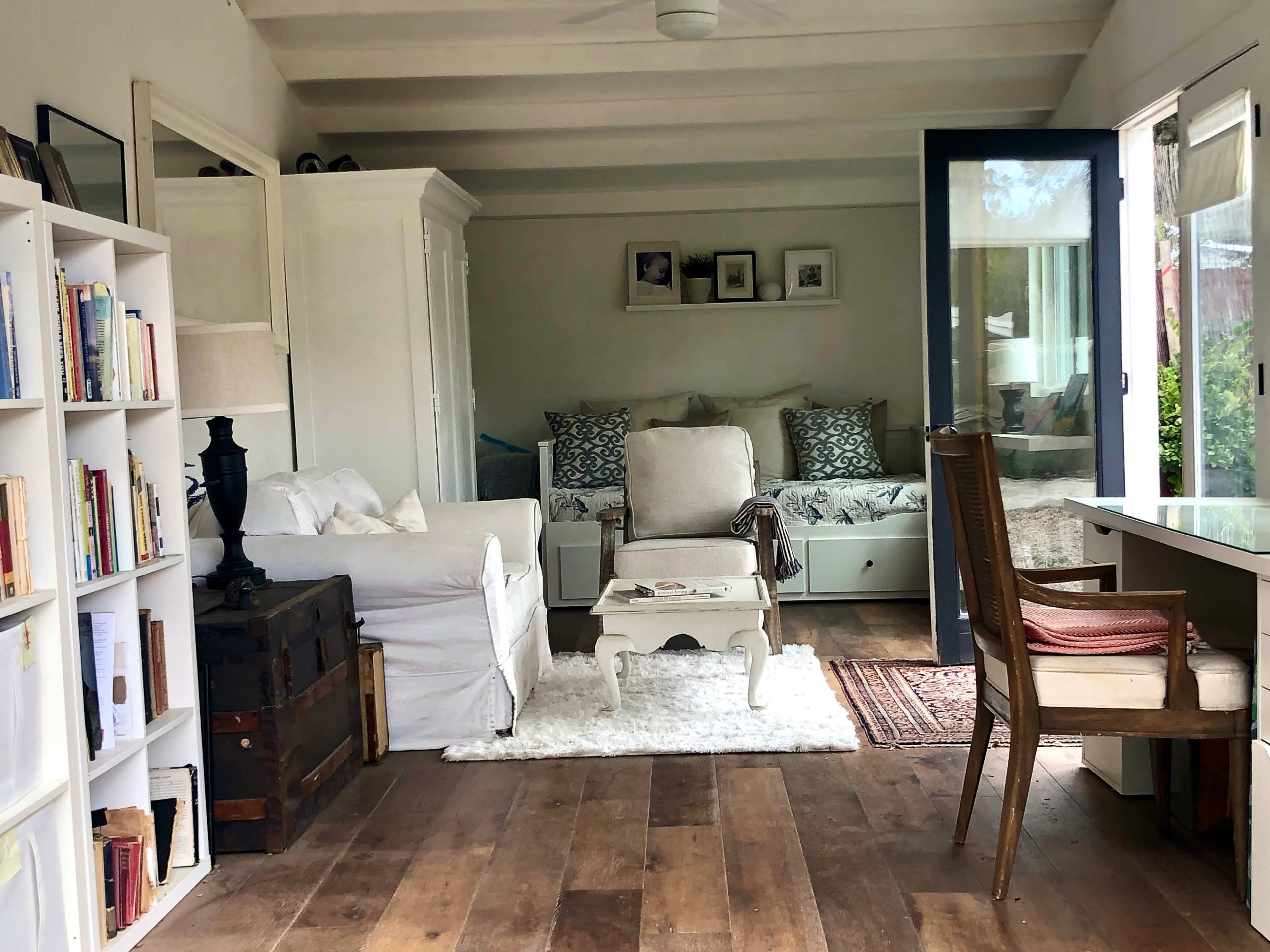 A cozy interior space featuring a living area with a white sofa, a small coffee table, a bookshelf, and a desk, all set against wooden flooring.