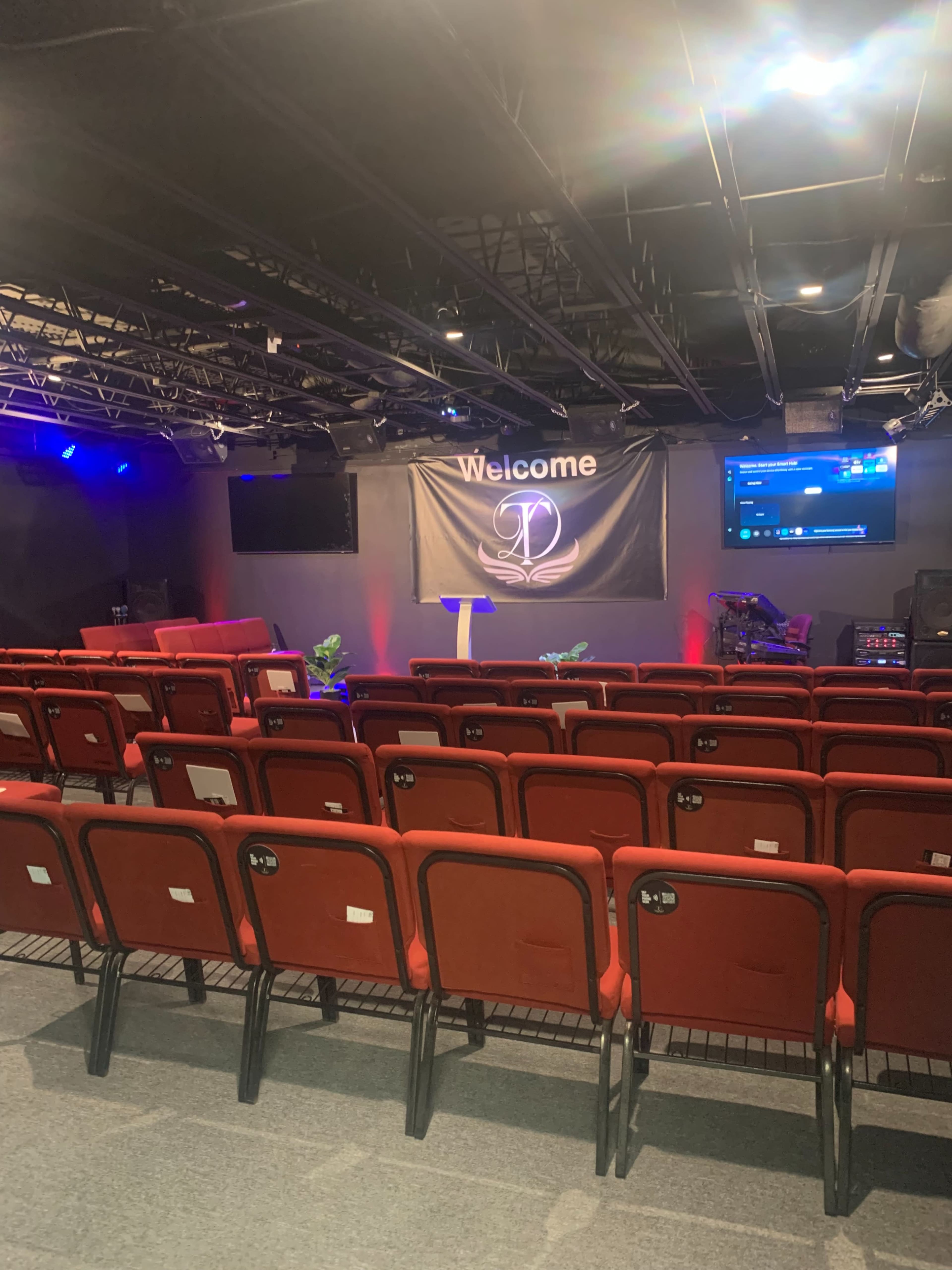 The image shows a small auditorium or theater with red chairs arranged in rows facing a stage, which features a table and backdrop with a sign that reads "Welcome."