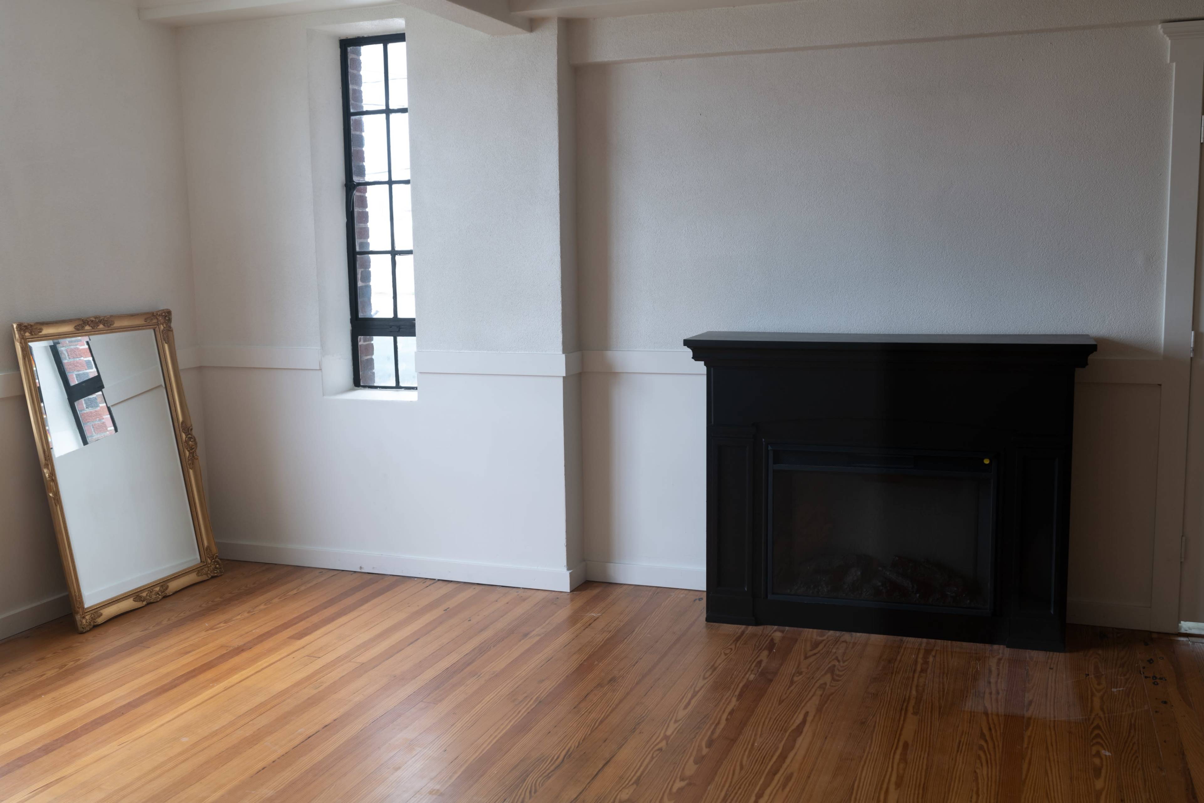 A sparsely furnished room with wooden flooring, featuring a black fireplace and a gold-framed mirror on the left wall.