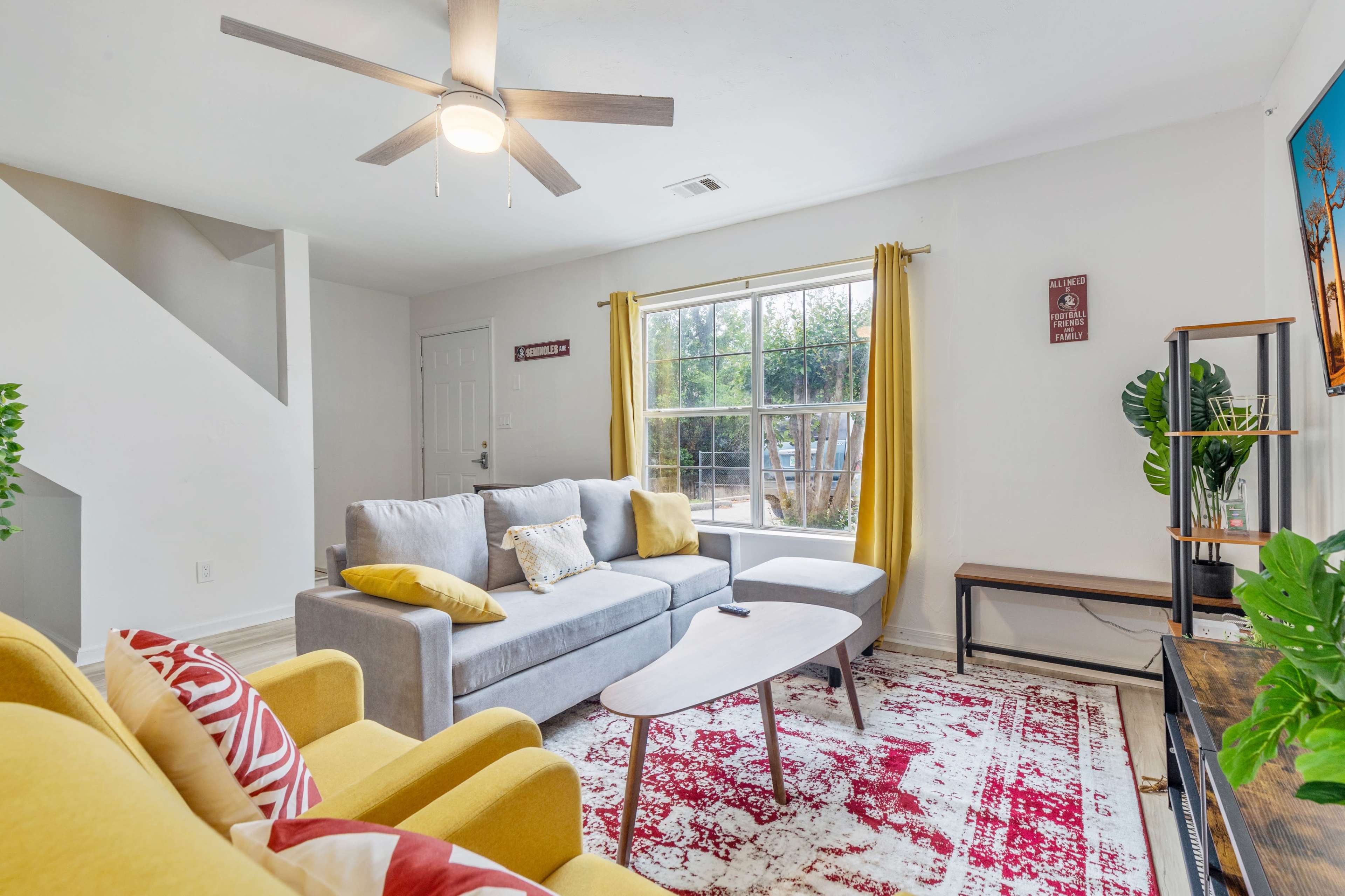 A bright living room features a gray sectional sofa, a yellow armchair, and a large window with sheer curtains, complemented by a colorful area rug and a wooden coffee table.
