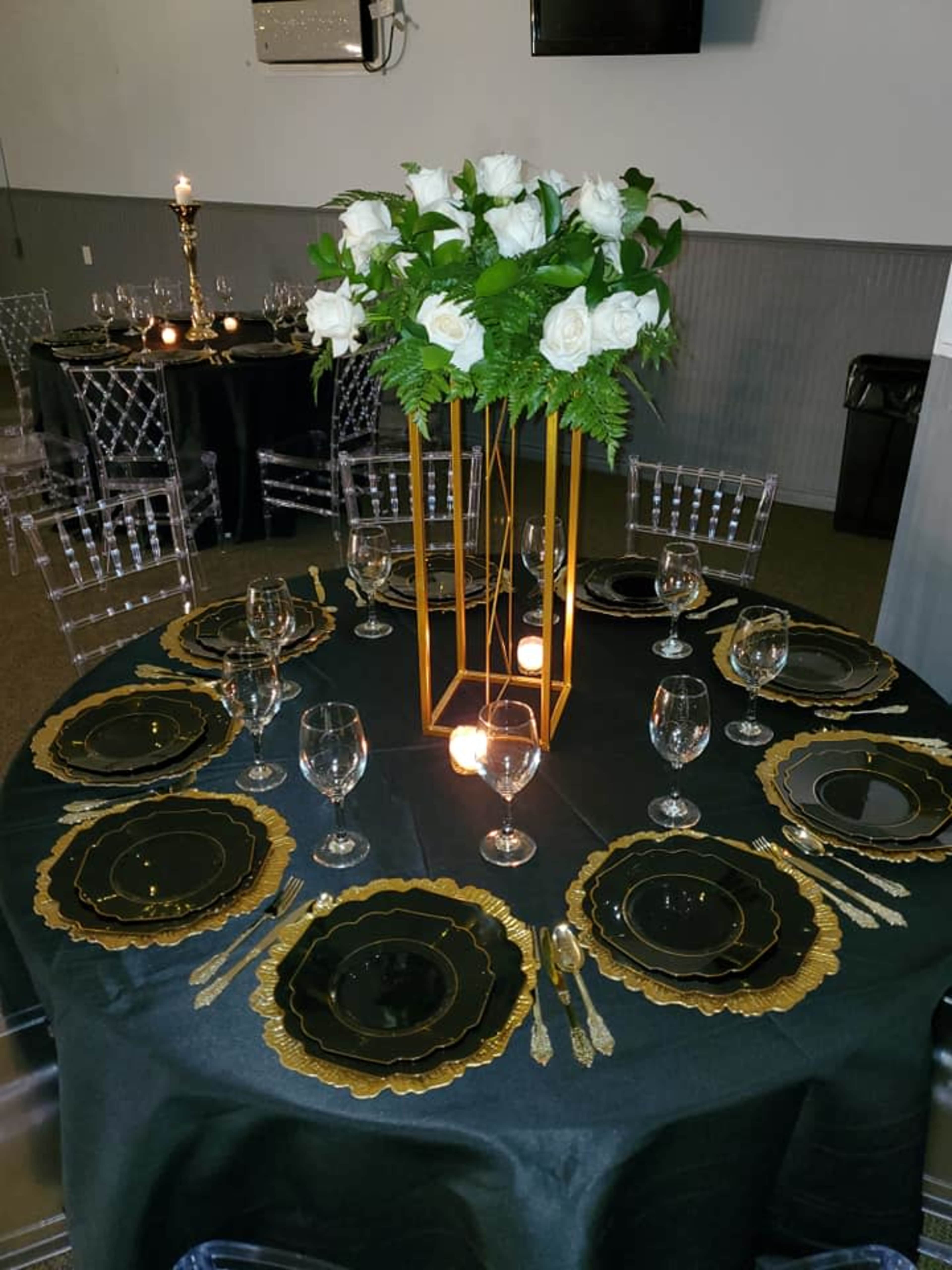 A dining table is elegantly set with black and gold tableware, featuring a tall floral centerpiece of white roses.