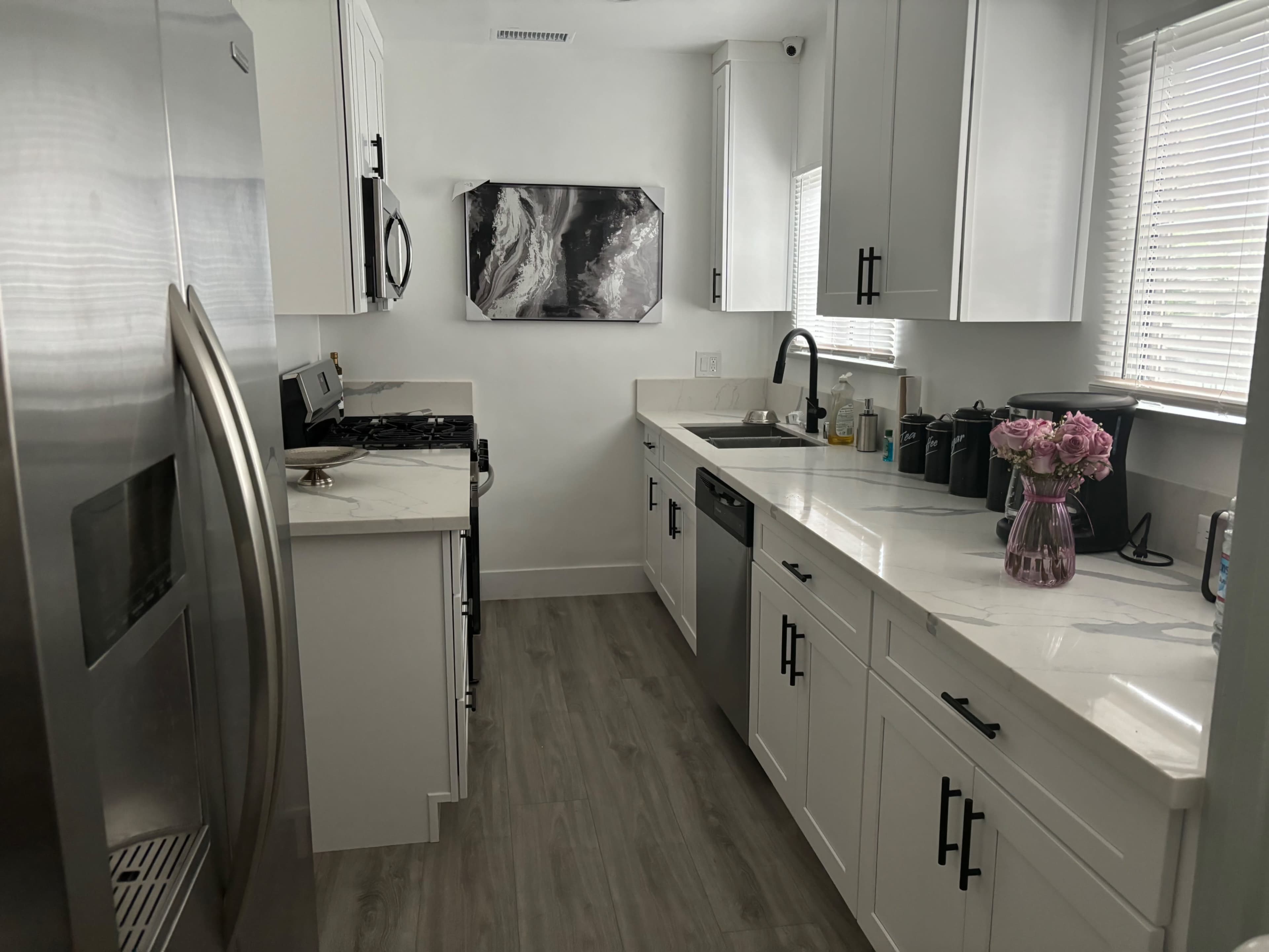 The image shows a modern kitchen featuring stainless steel appliances, white cabinetry, and a marble countertop with a vase of flowers on the counter.