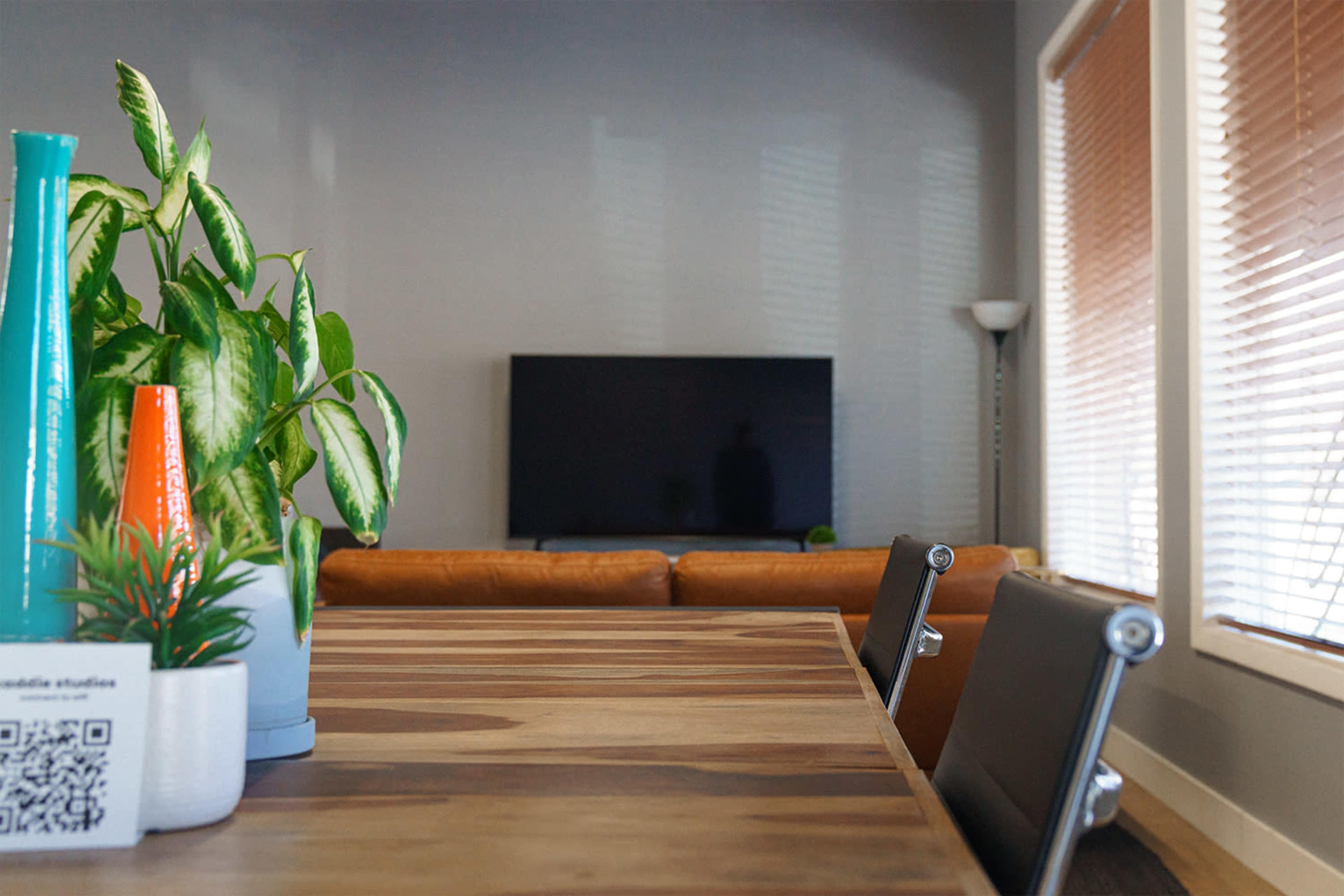 A wooden dining table with a plant centerpiece is shown in a room featuring a brown sofa and a television.