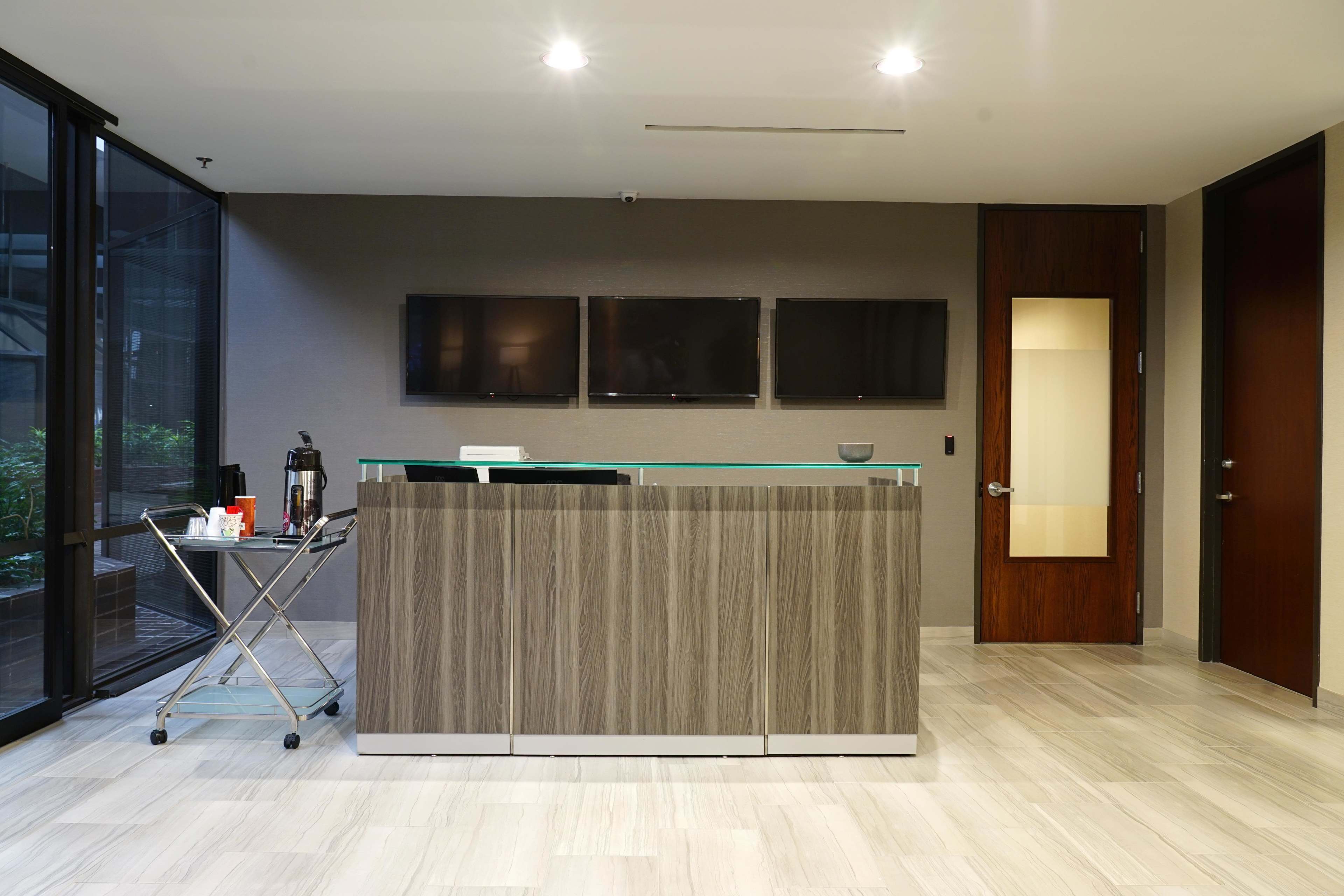 A modern reception area with a wooden front desk, three wall-mounted televisions, and a side cart next to a glass wall.