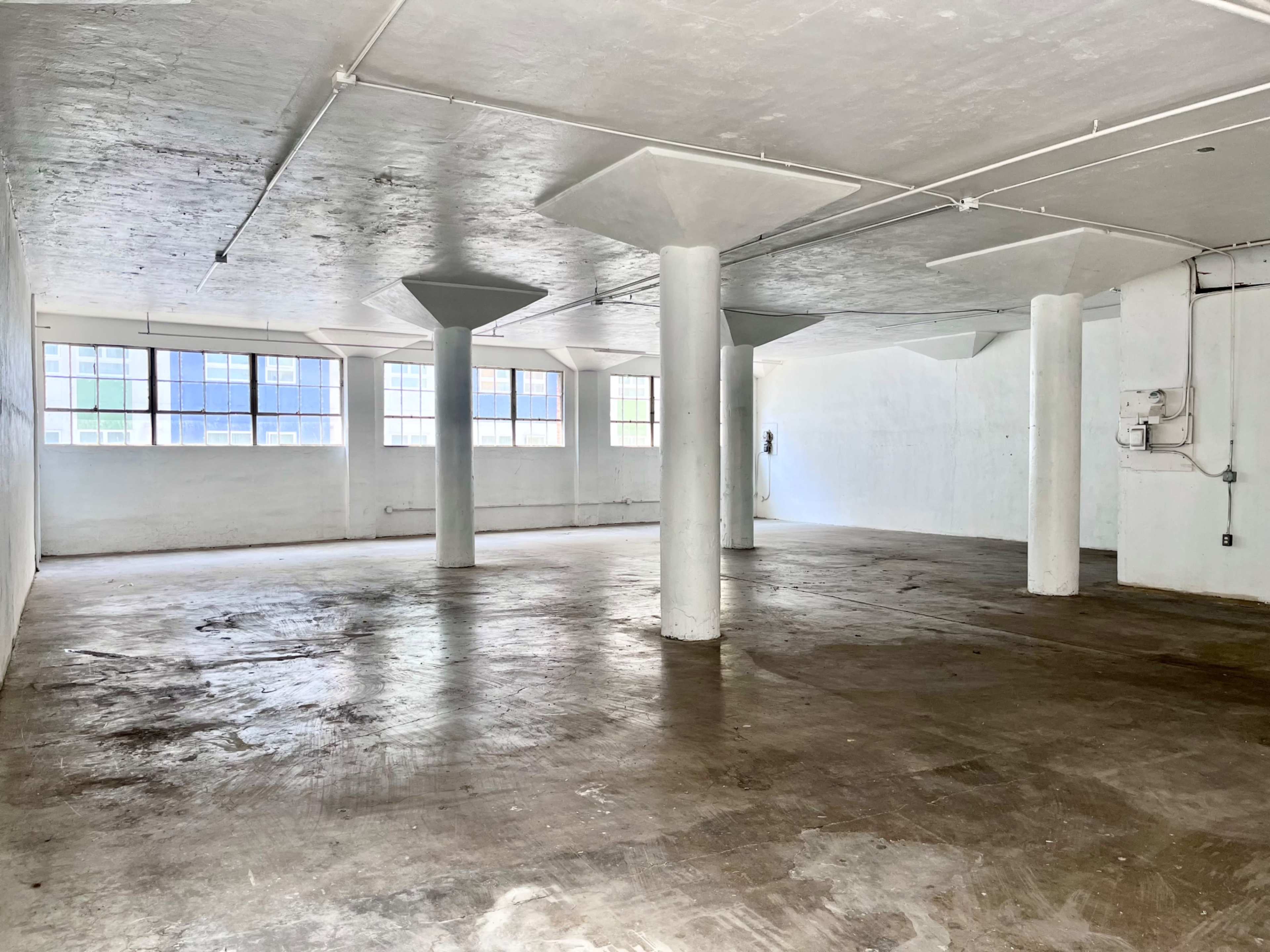 A large, empty interior space with concrete floors, white walls, and several columns, illuminated by natural light from multiple windows.