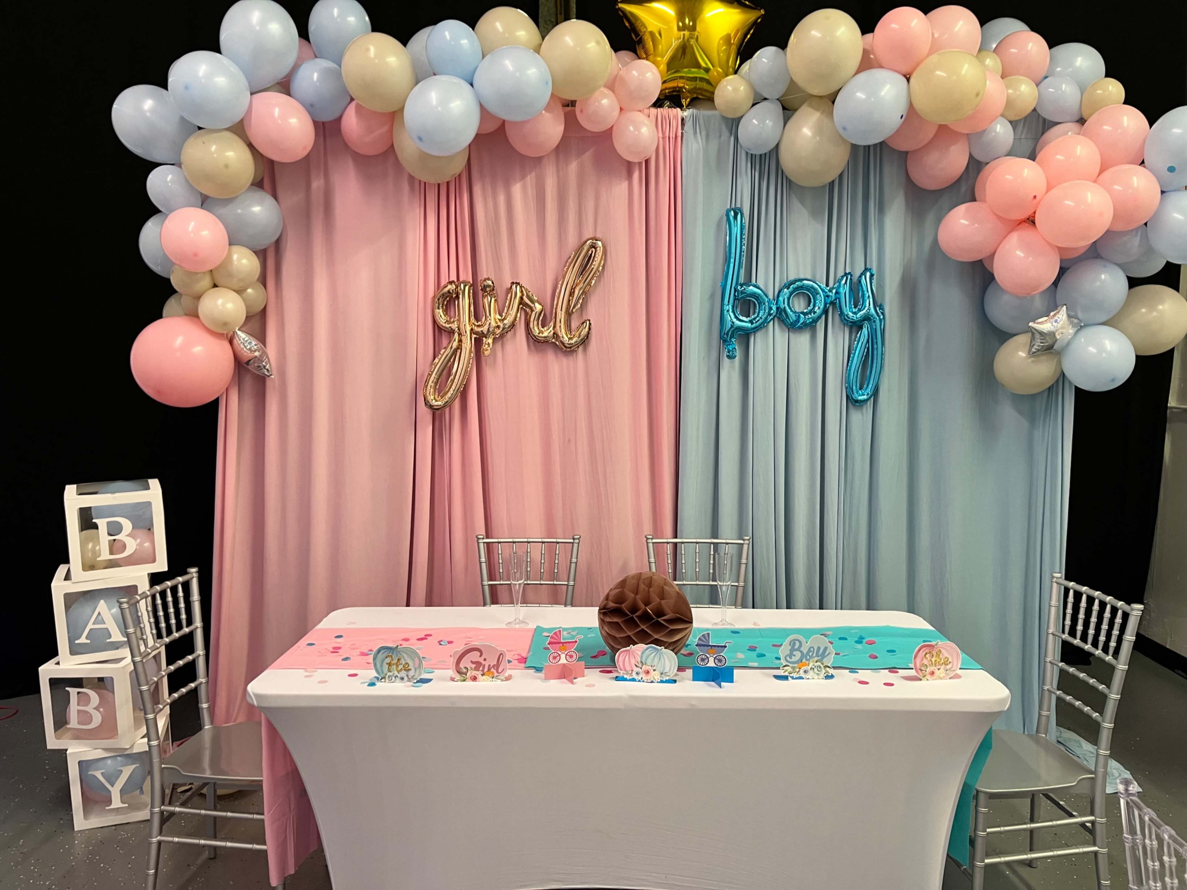 The image shows a decorated table for a baby shower, featuring pink and blue balloons, a "girl" and "boy" sign, and a table draped with pastel colors.