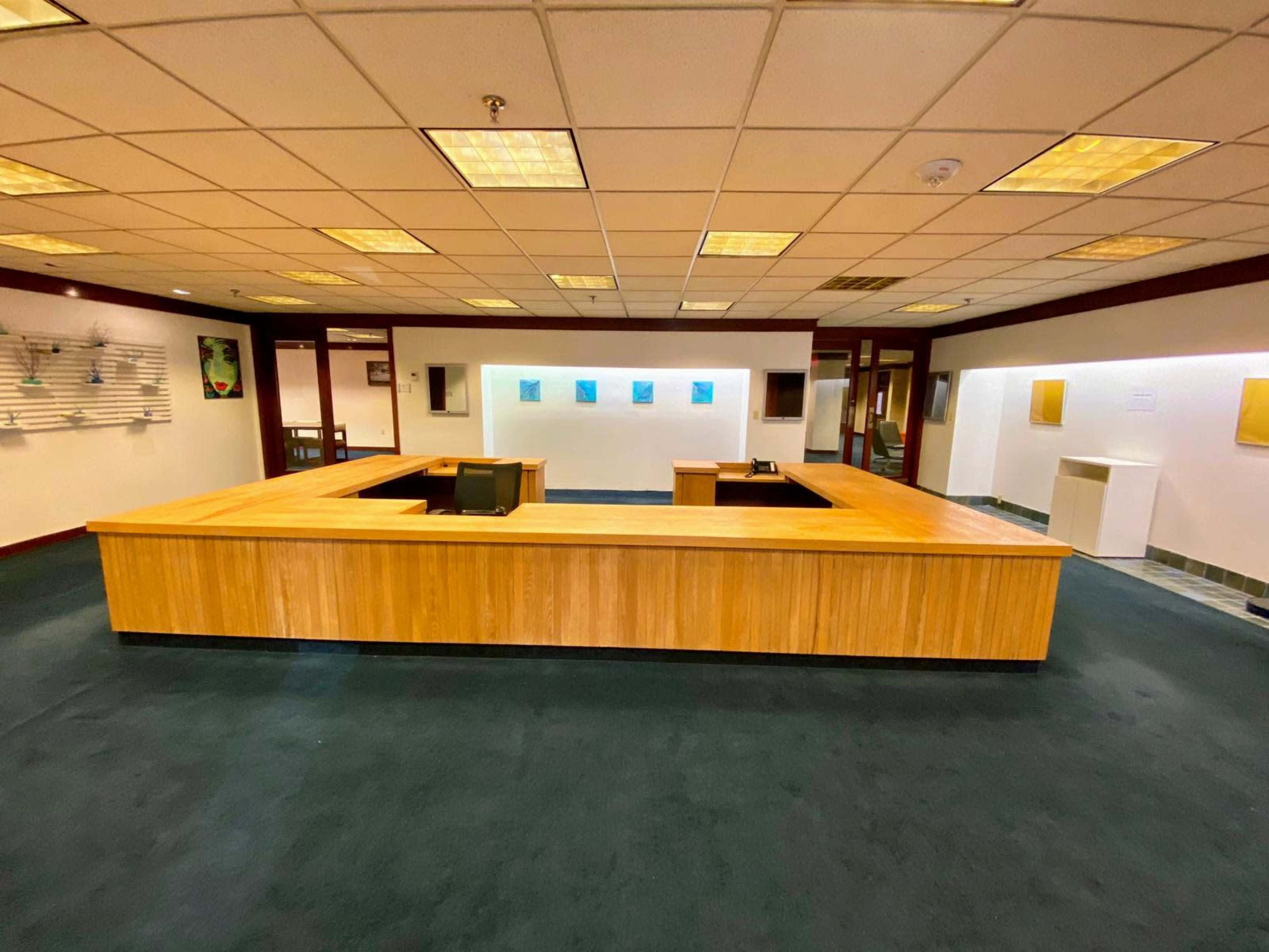 The image shows a spacious office reception area with a large wooden reception desk at its center, surrounded by green carpet and white walls adorned with artwork.