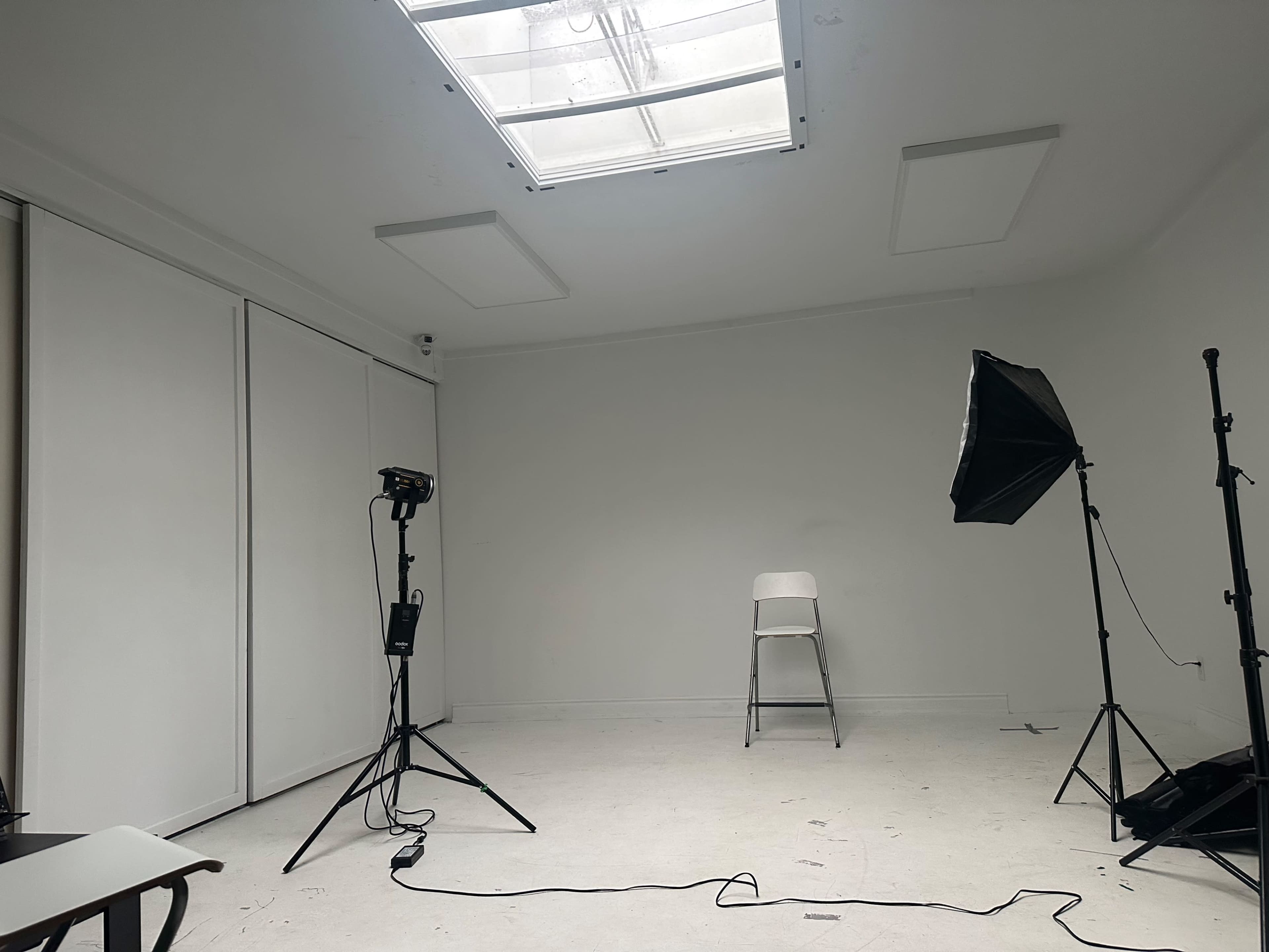The image shows a minimalistic studio room with a single white chair, a softbox light, and a backdrop setup under a skylight.