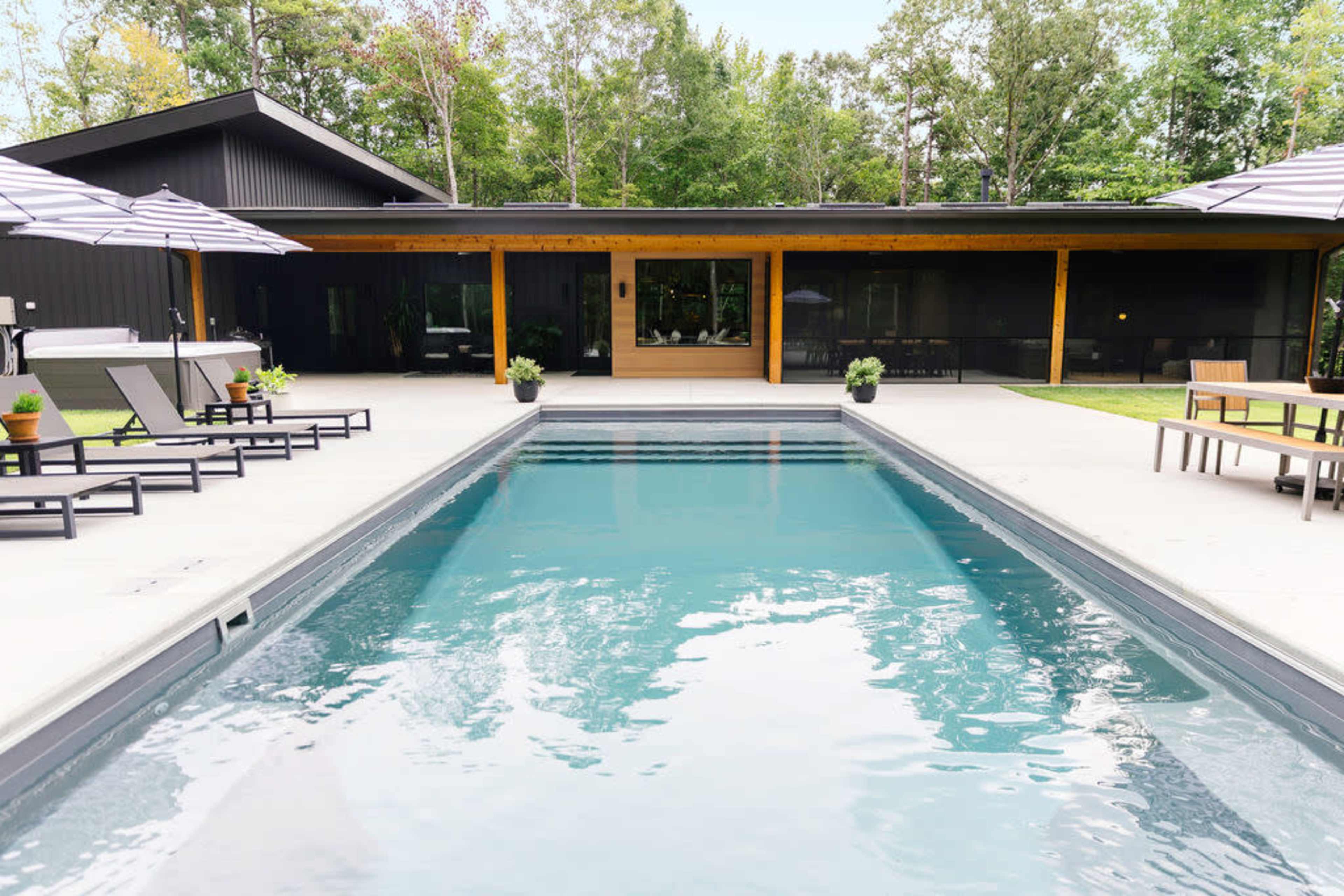 A modern outdoor area features a swimming pool in front of a sleek black house surrounded by trees and patio furniture.
