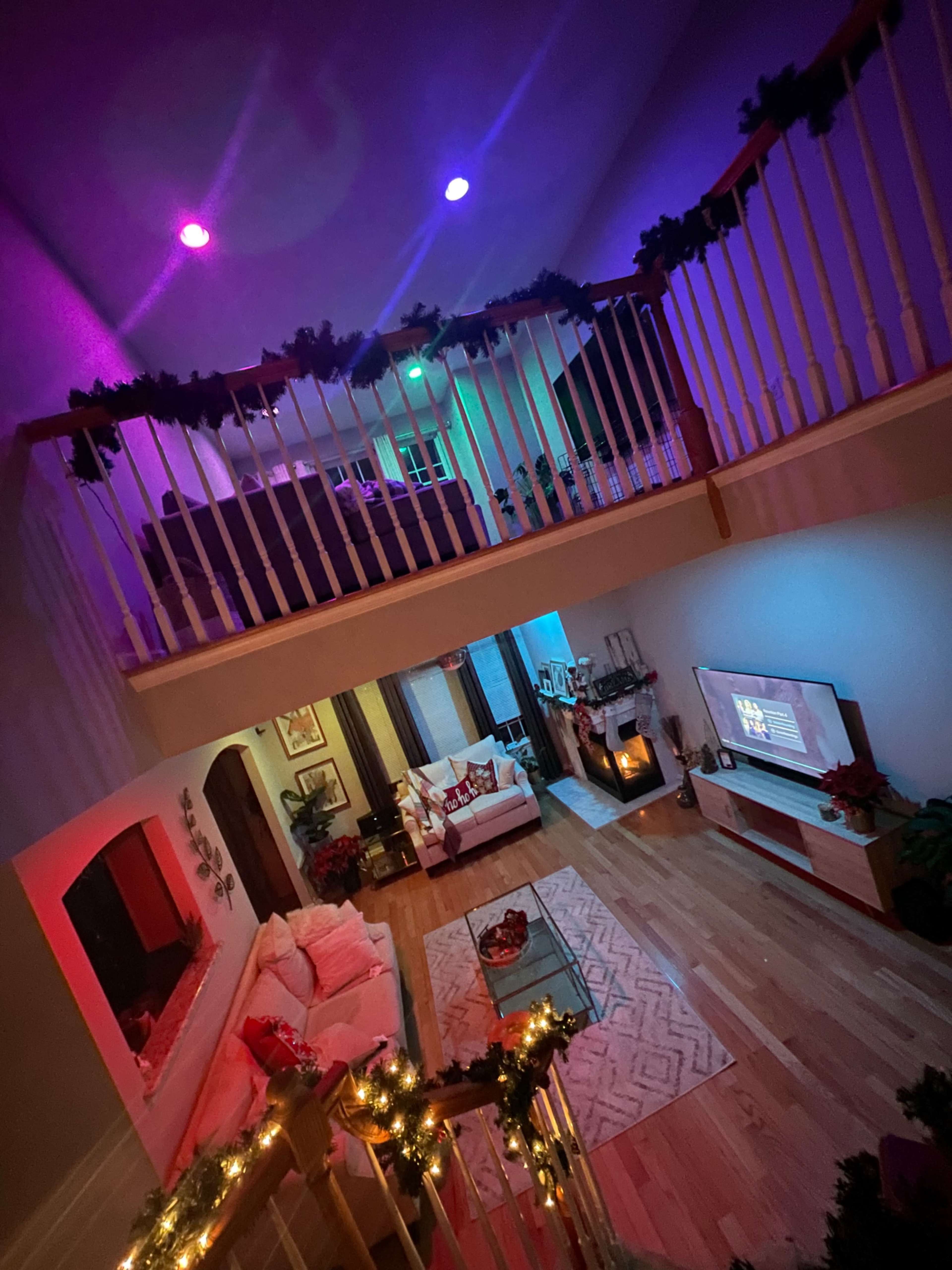 A two-story living room decorated with colorful lights and garlands, featuring a modern sofa, a television, and a fireplace.