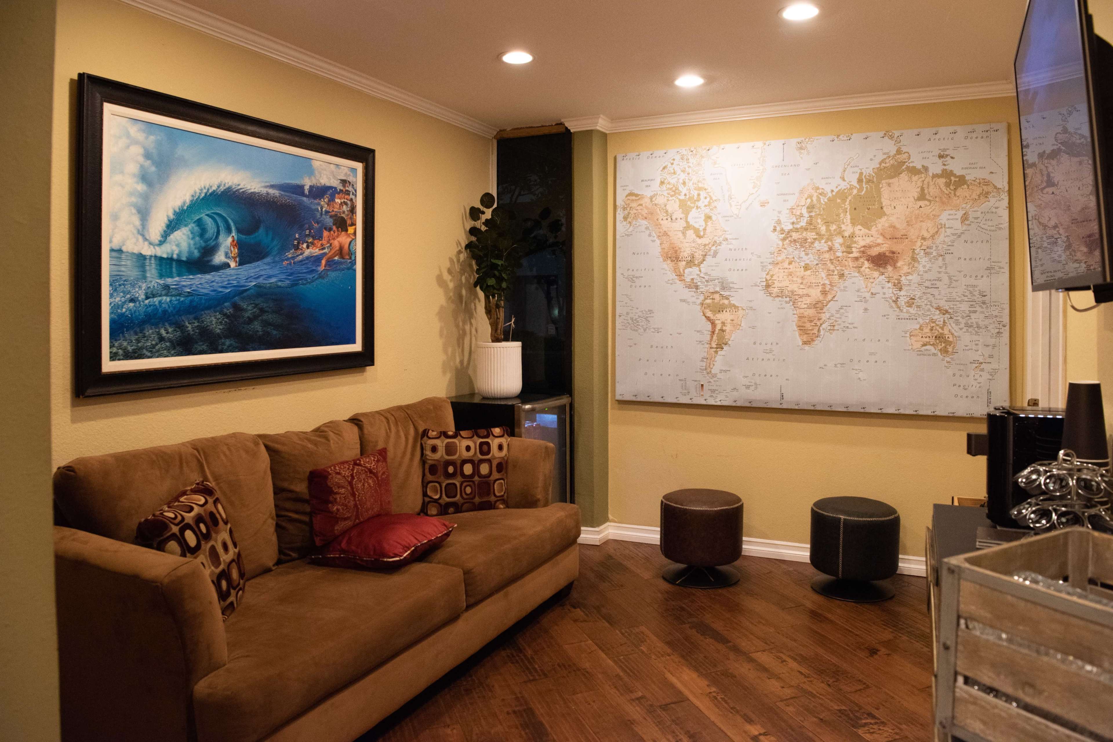 The image shows a cozy living room featuring a brown couch with decorative pillows, a large framed wave painting on one wall, and a world map mounted on another wall.