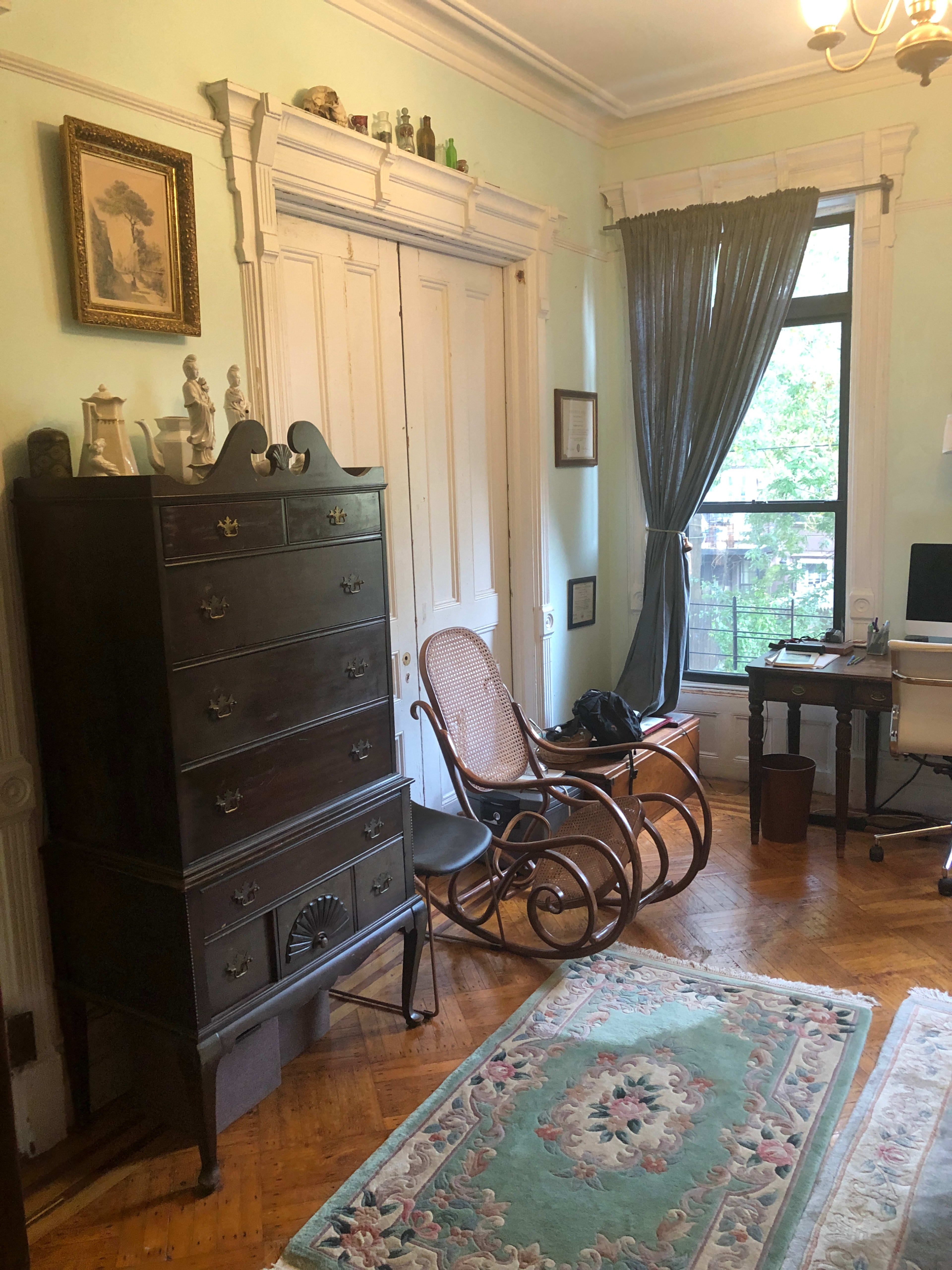 A room with a wooden dresser, a rocking chair, a small desk, and a window draped with a gray curtain.