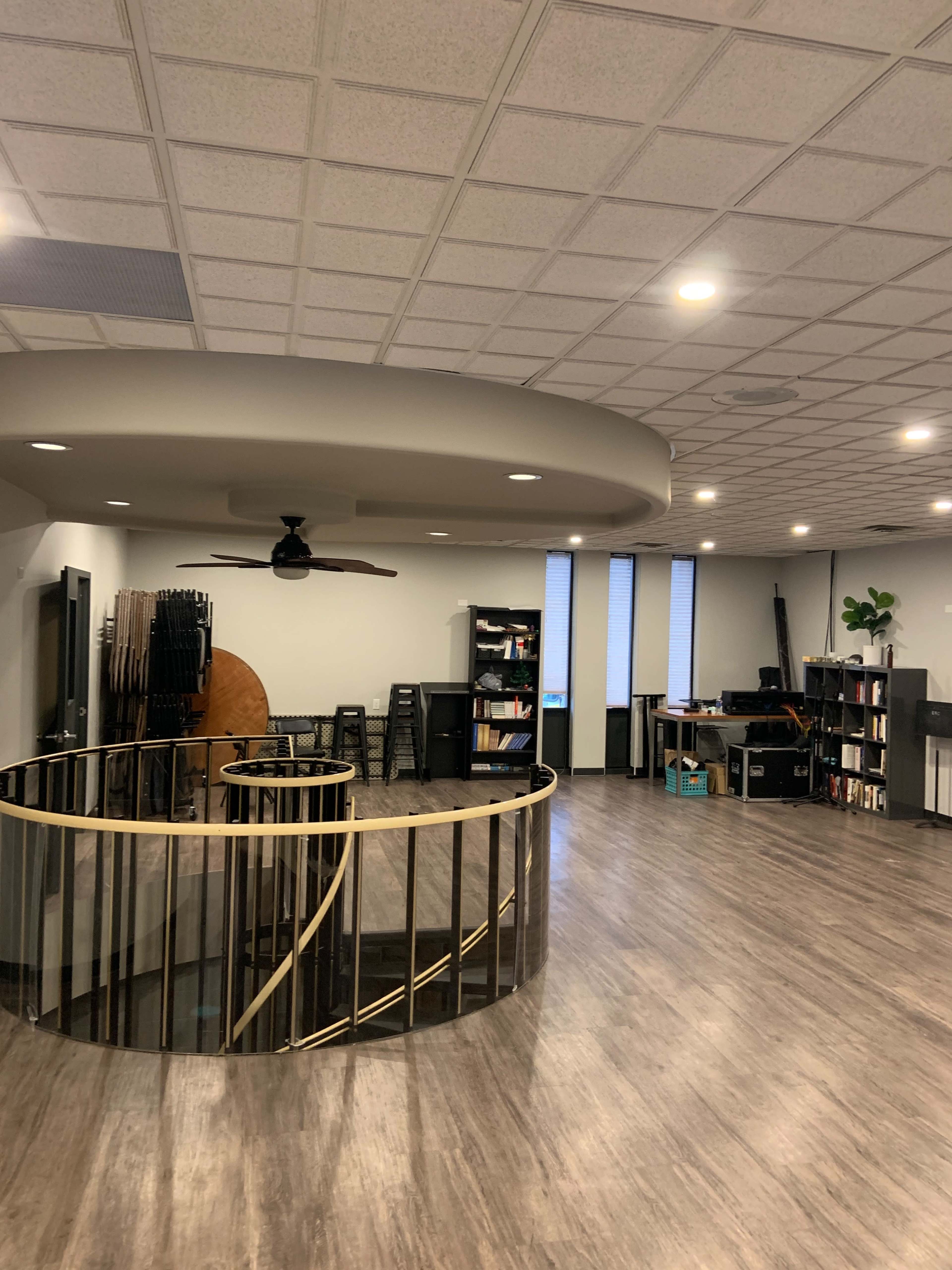 The image shows a spacious, modern interior with a circular railing, a ceiling fan, and bookshelves against the walls.