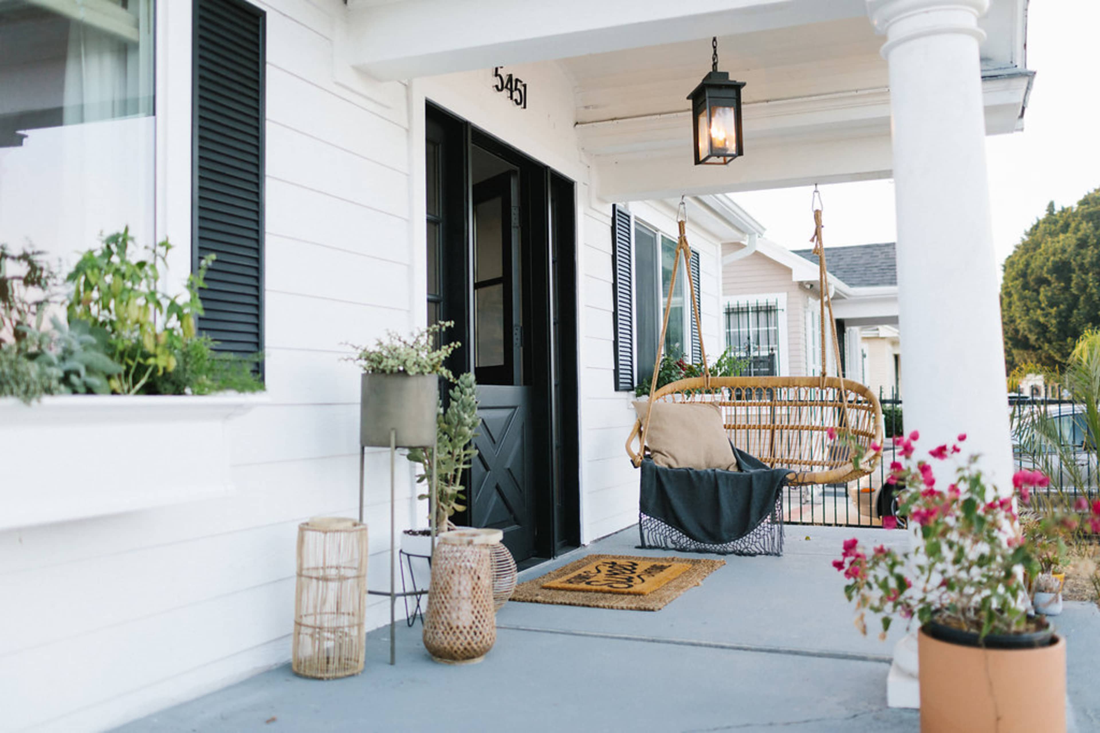 A front porch features a swing chair, potted plants, and a welcome mat beside a black door.
