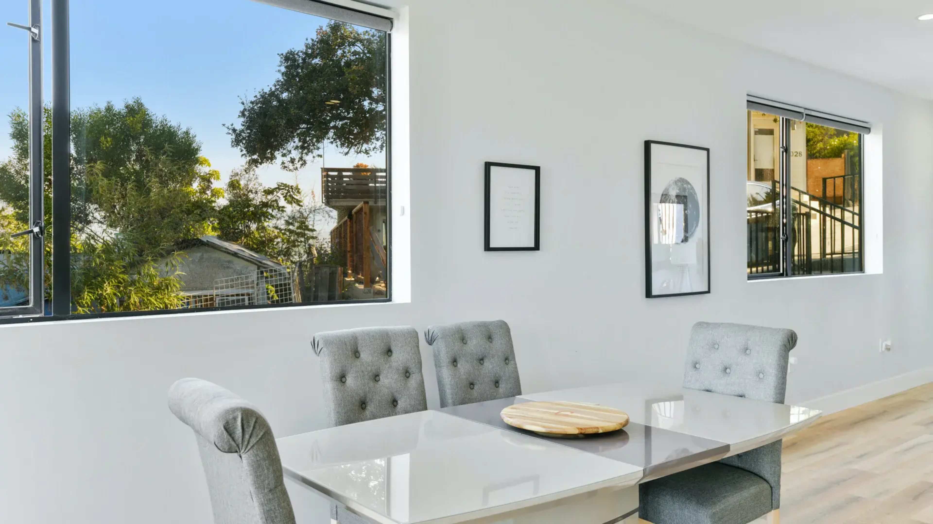 A modern dining area features a glass table with tufted gray chairs and large windows revealing greenery outside.