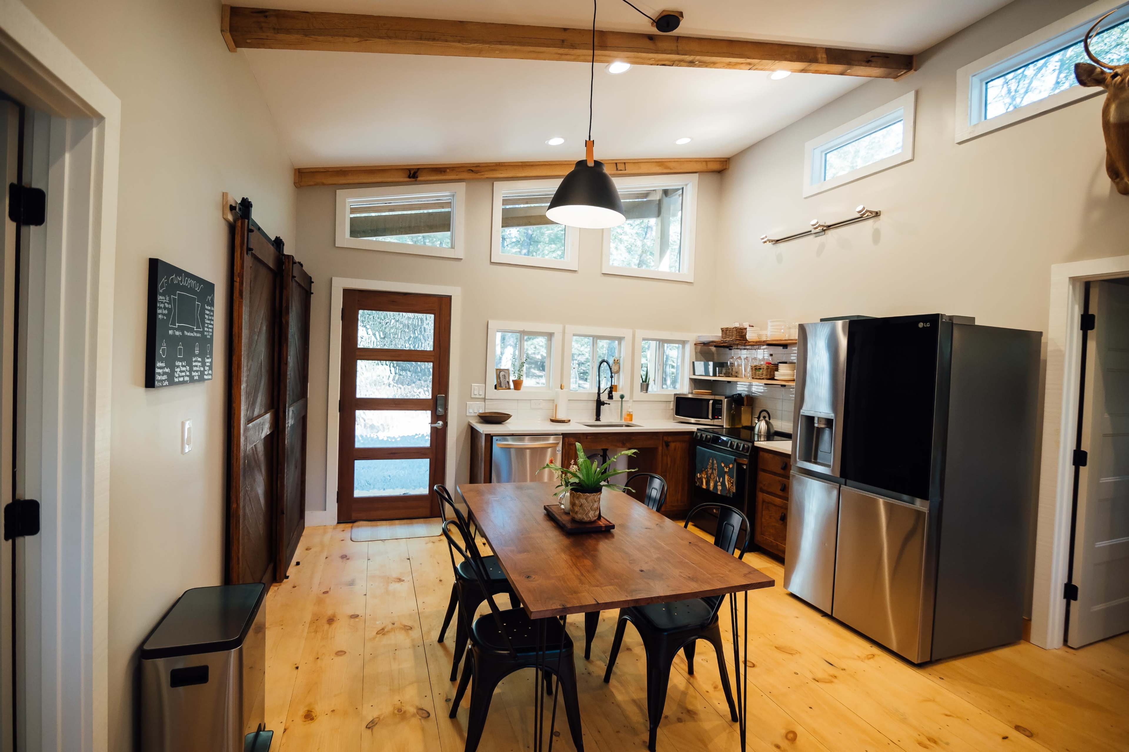 A modern kitchen with a wooden dining table, stainless steel refrigerator, and a large window allowing natural light to enter.