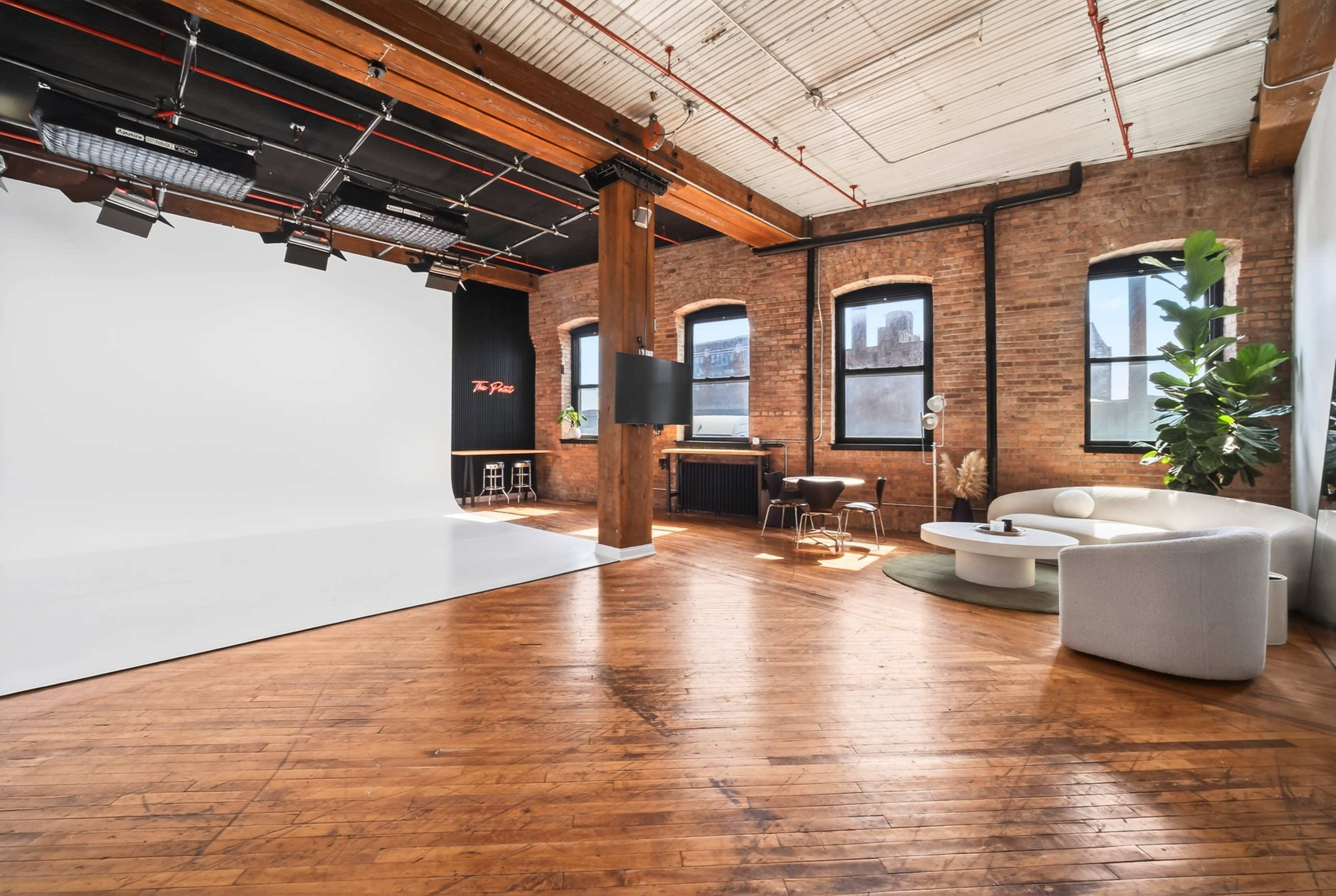 The image shows a spacious, well-lit studio with wooden floors, exposed brick walls, a large white backdrop, and modern furniture.