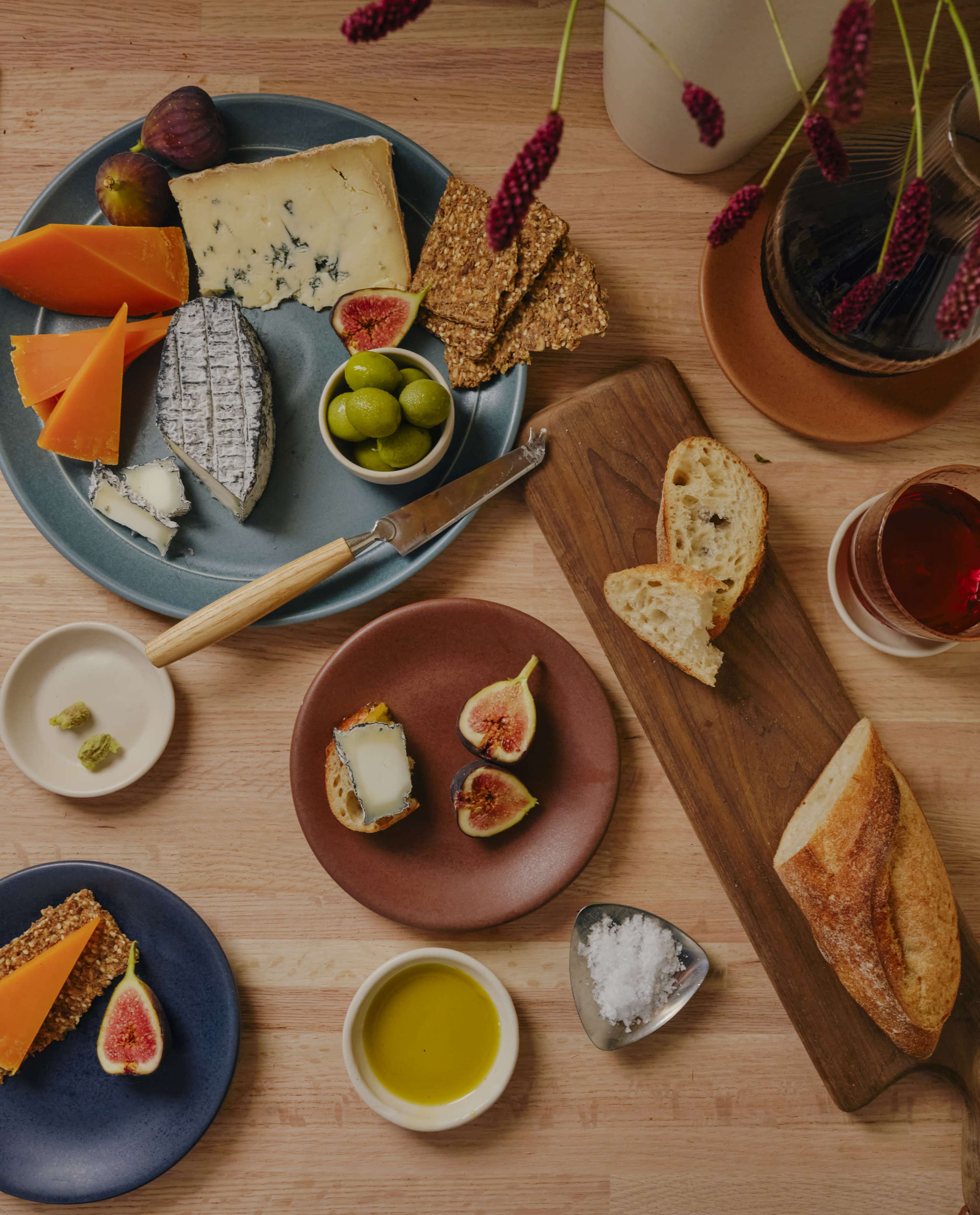 A wooden table displays a variety of cheeses, olives, figs, bread, and olive oil, arranged on colorful plates and boards alongside a glass of red beverage.