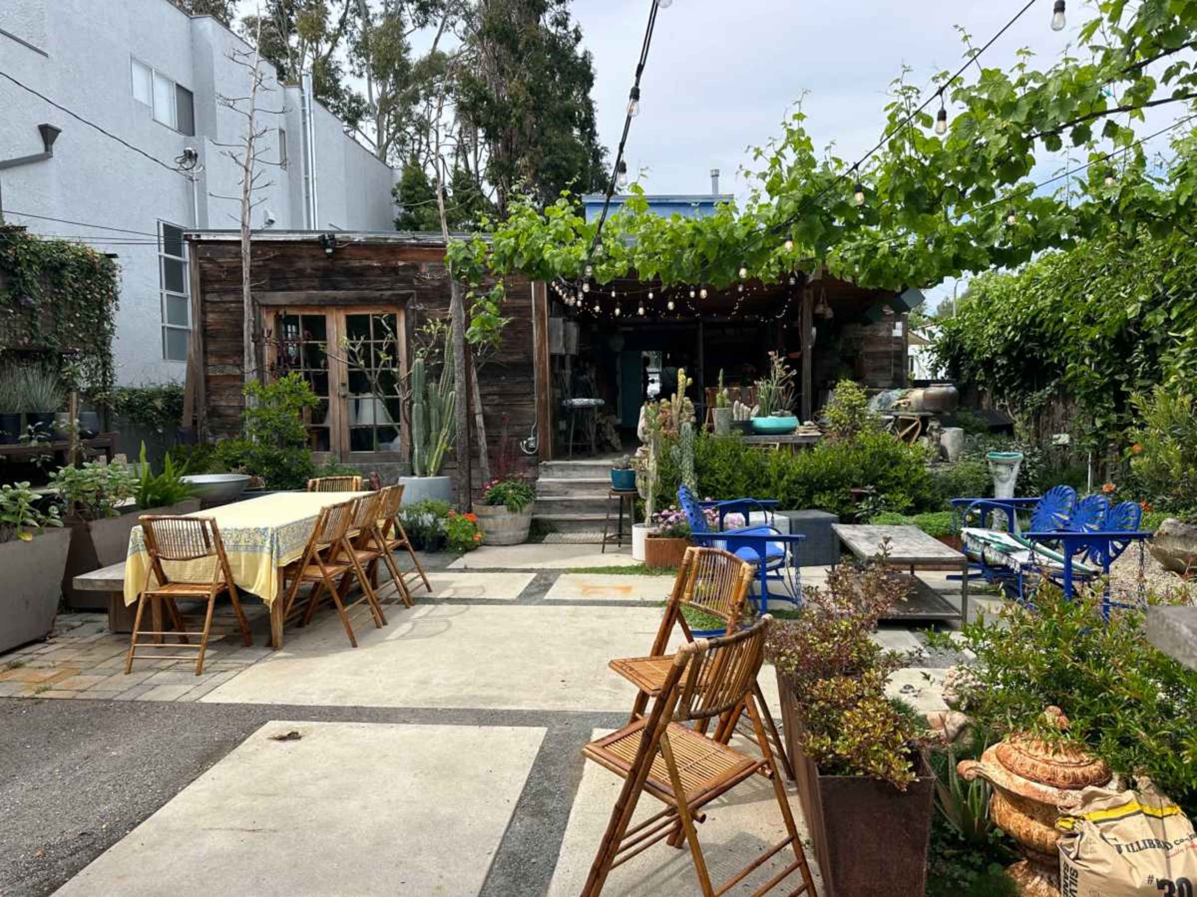The image shows an outdoor patio with various seating arrangements, a dining table, greenery, and a rustic building in the background.