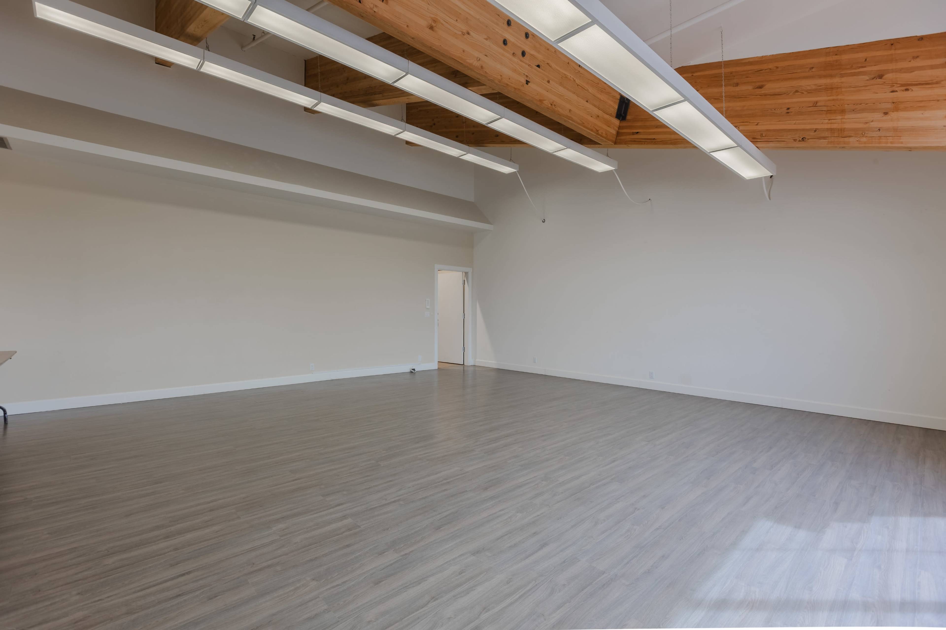 The image depicts a spacious, empty room with wooden beams and overhead lighting.