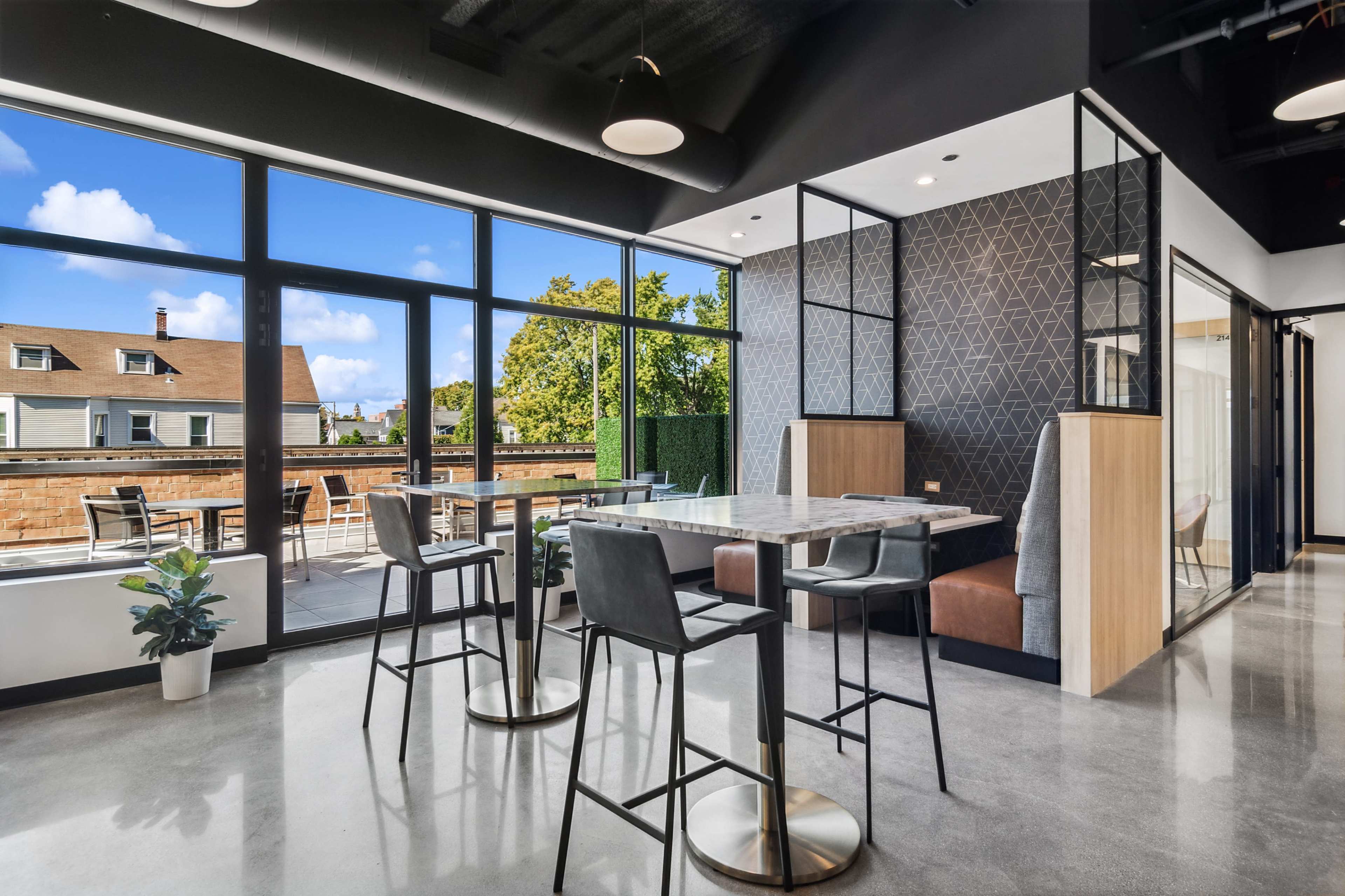 The image shows a modern office space featuring a dining area with high tables and stools, large windows revealing an outdoor terrace, and geometric wall designs.