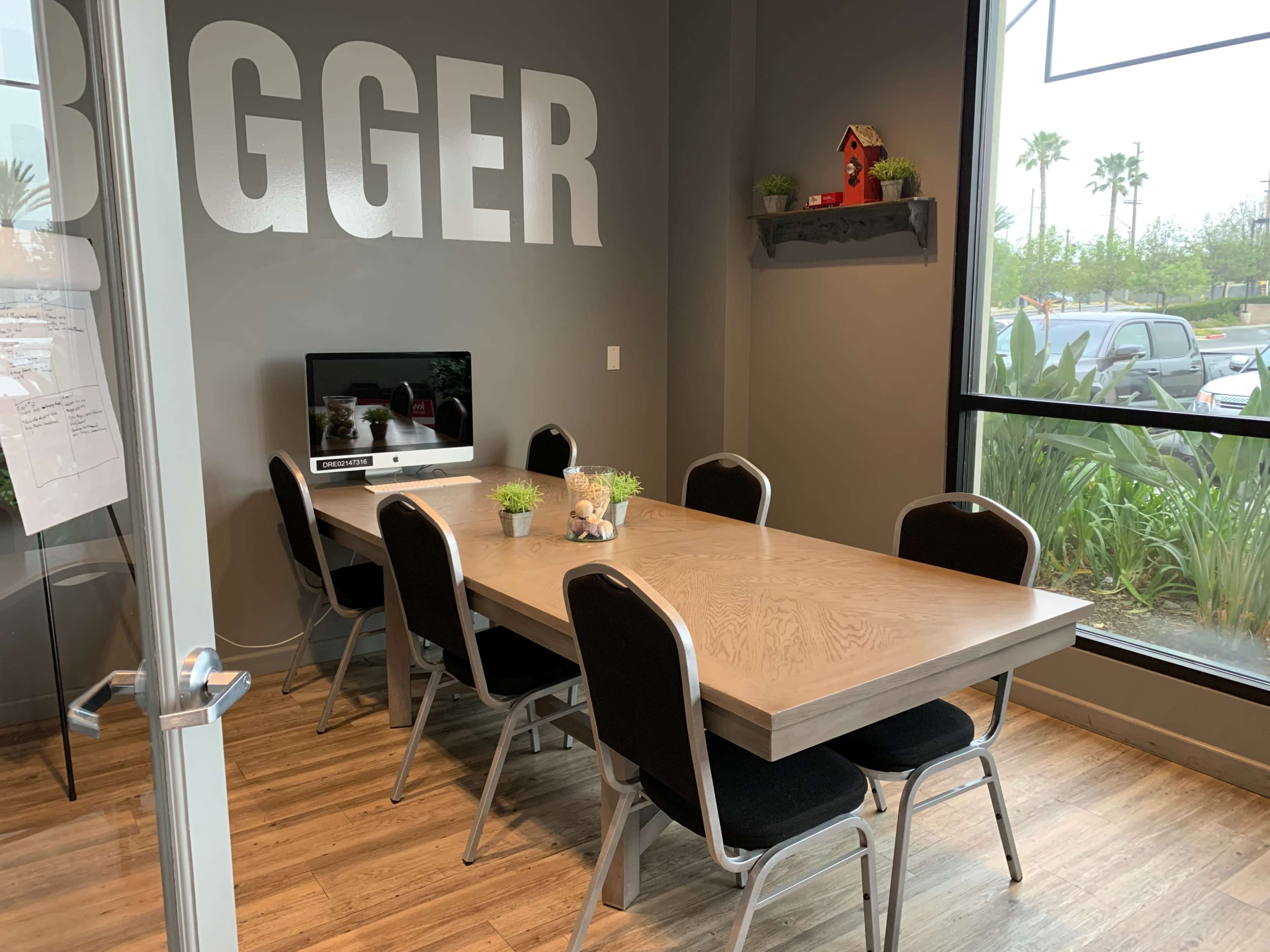 A conference room features a long wooden table surrounded by eight black chairs, with a television mounted on the wall and plants on a shelf.