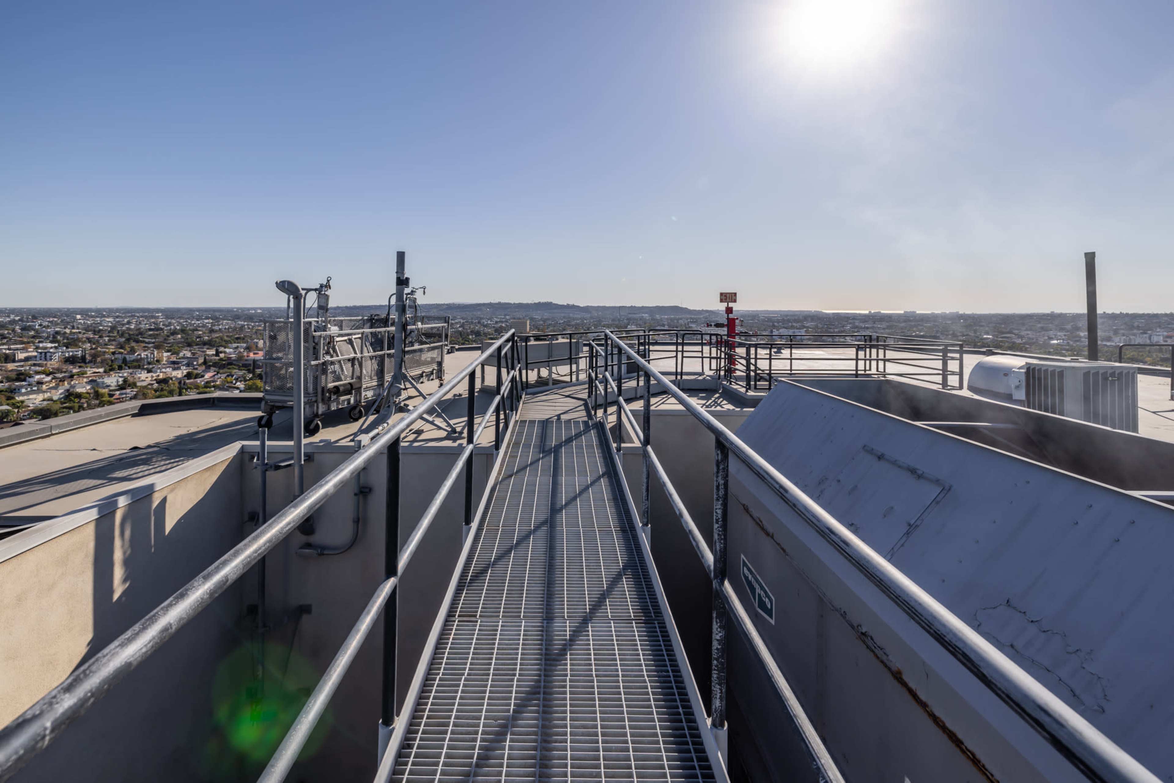 The image shows a metal walkway on a rooftop overlooking a vast urban landscape beneath a clear sky.