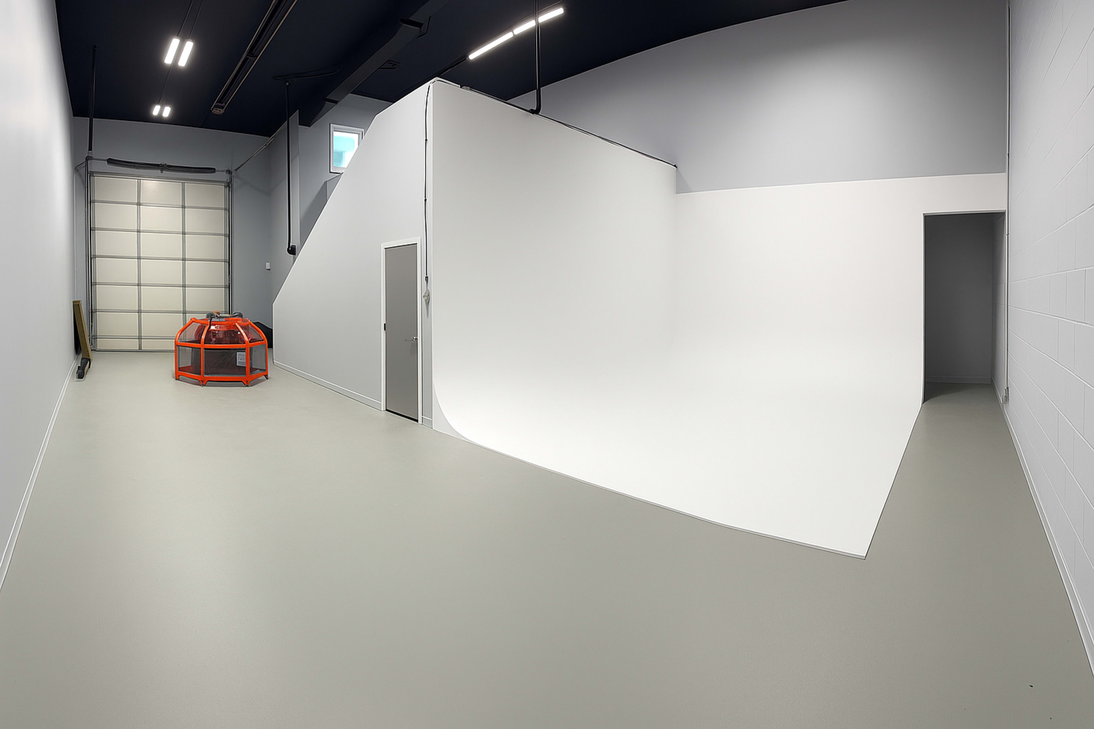 The image shows a spacious, well-lit photography studio featuring a curved white backdrop, a gray floor, and a door leading to another room.