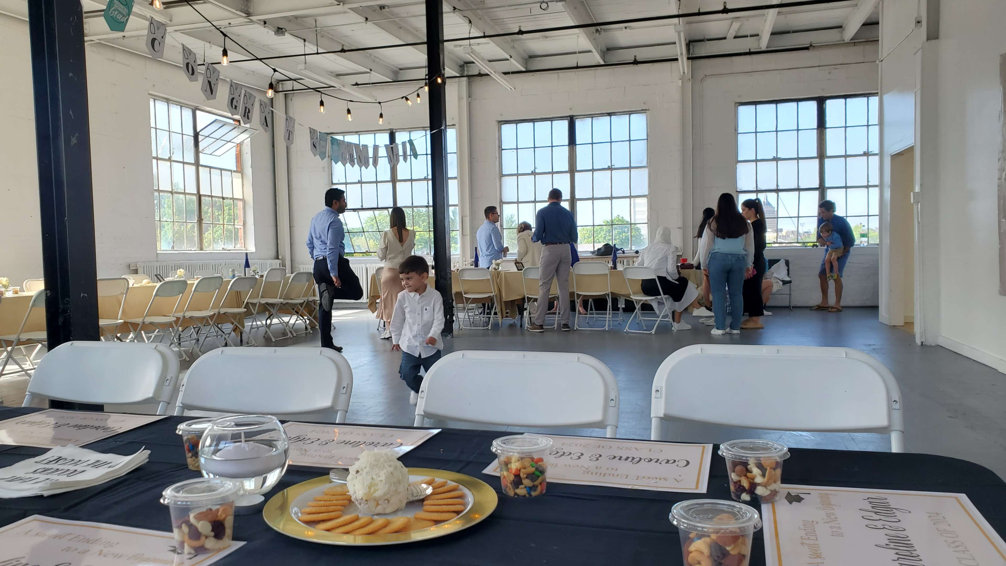 A spacious event hall features tables set for a gathering, with guests mingling near large windows that let in natural light.