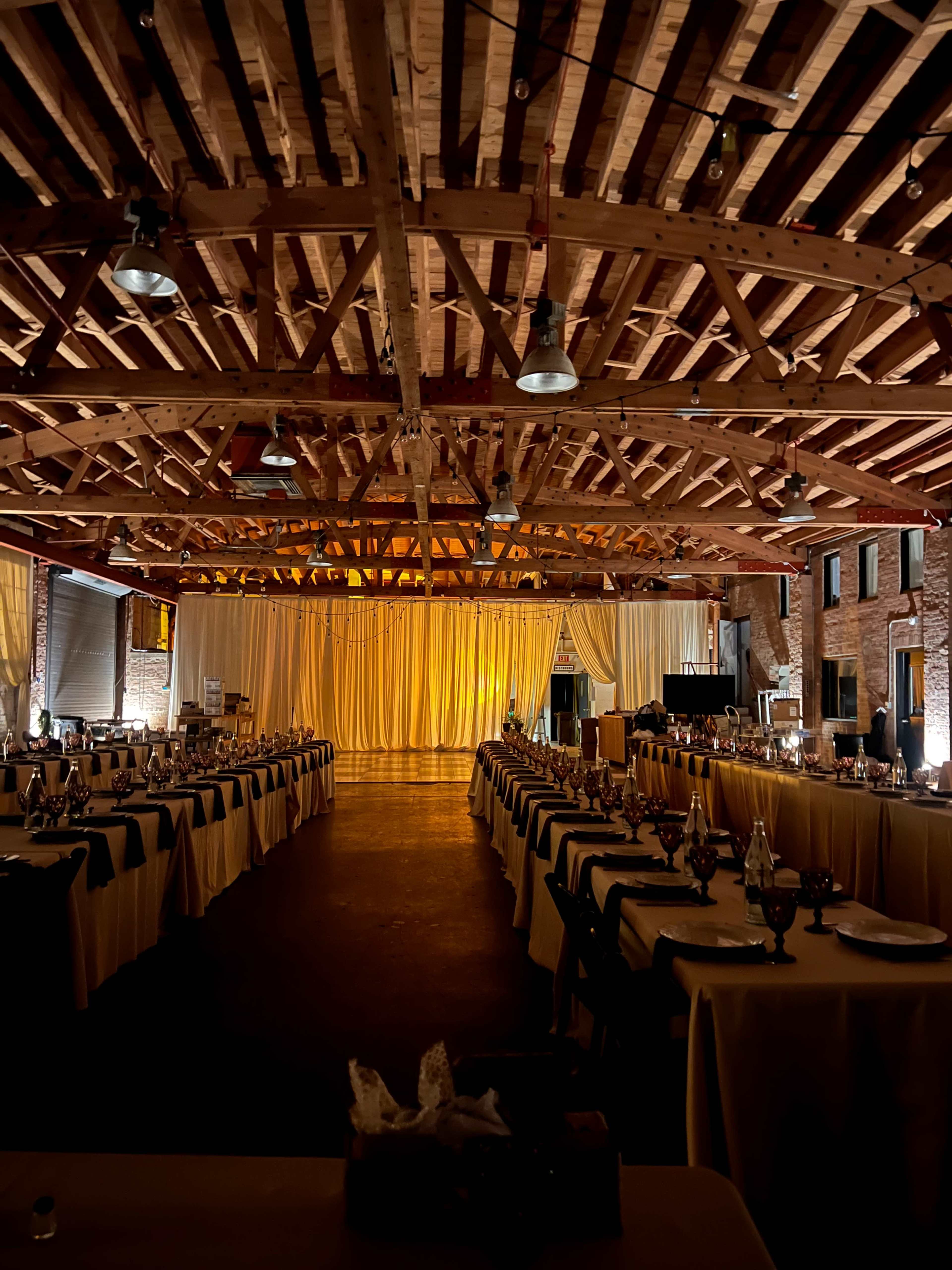 A spacious banquet hall features rows of elegantly set tables under a wooden beam ceiling, illuminated by warm lighting.