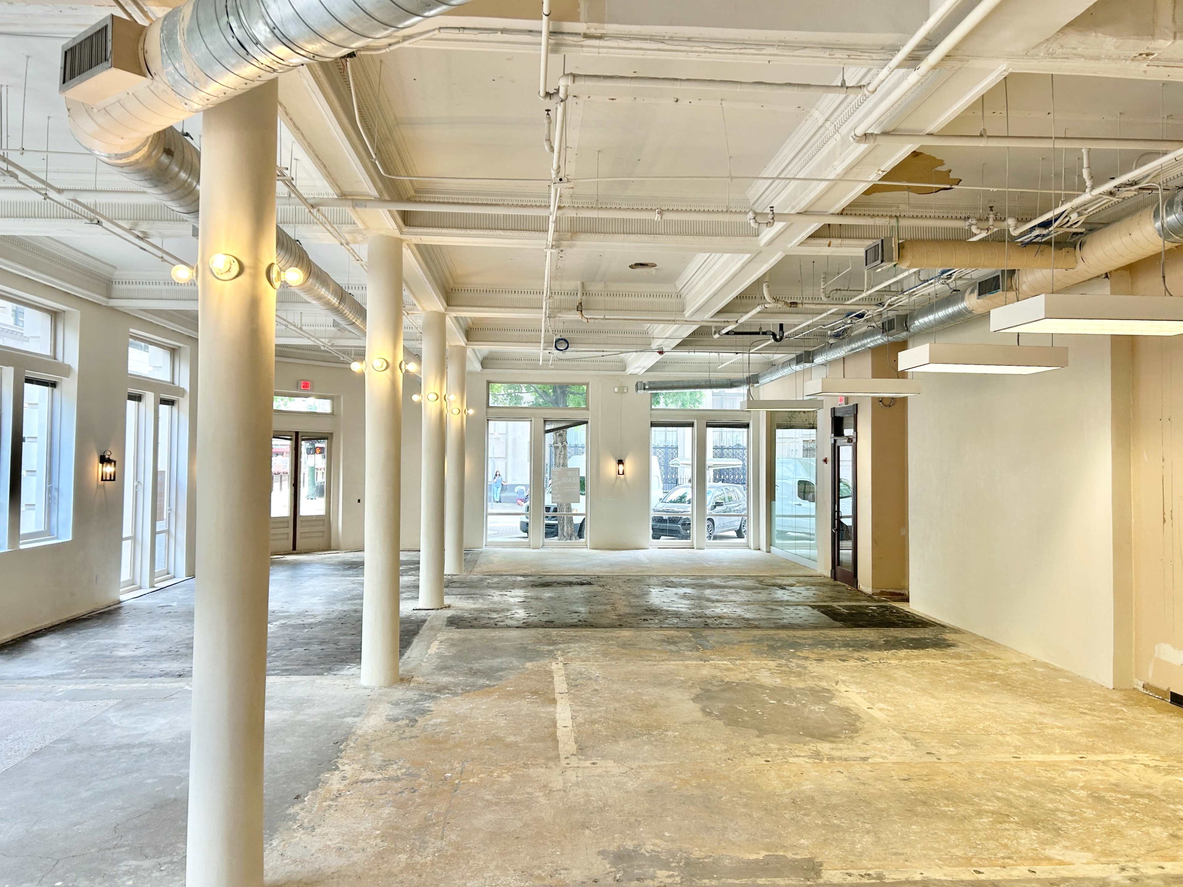 The image shows an empty commercial space with concrete floors, exposed pipes, and large windows allowing natural light to enter.