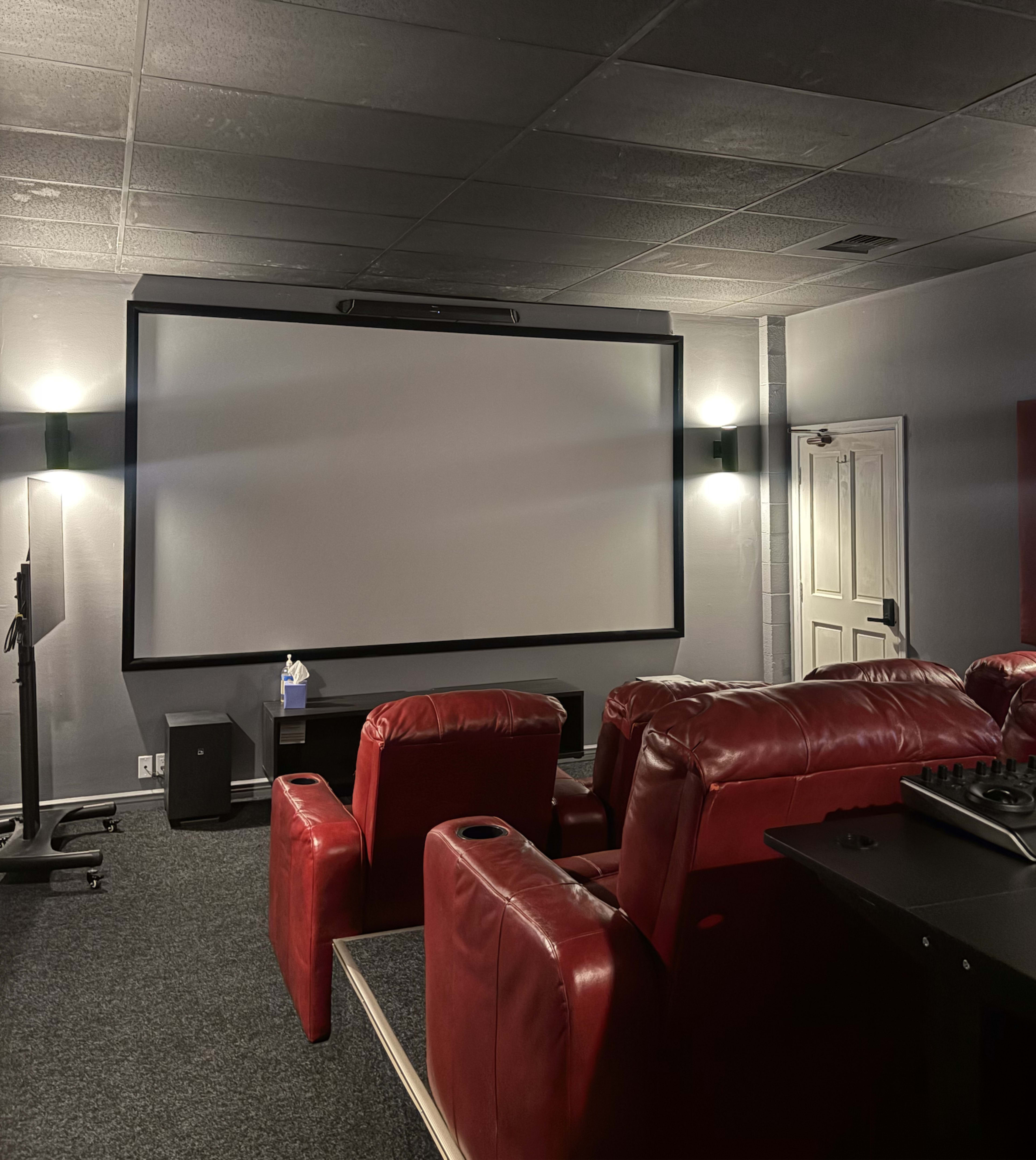The image shows a dimly lit home theater with red leather recliners facing a large, blank projection screen.